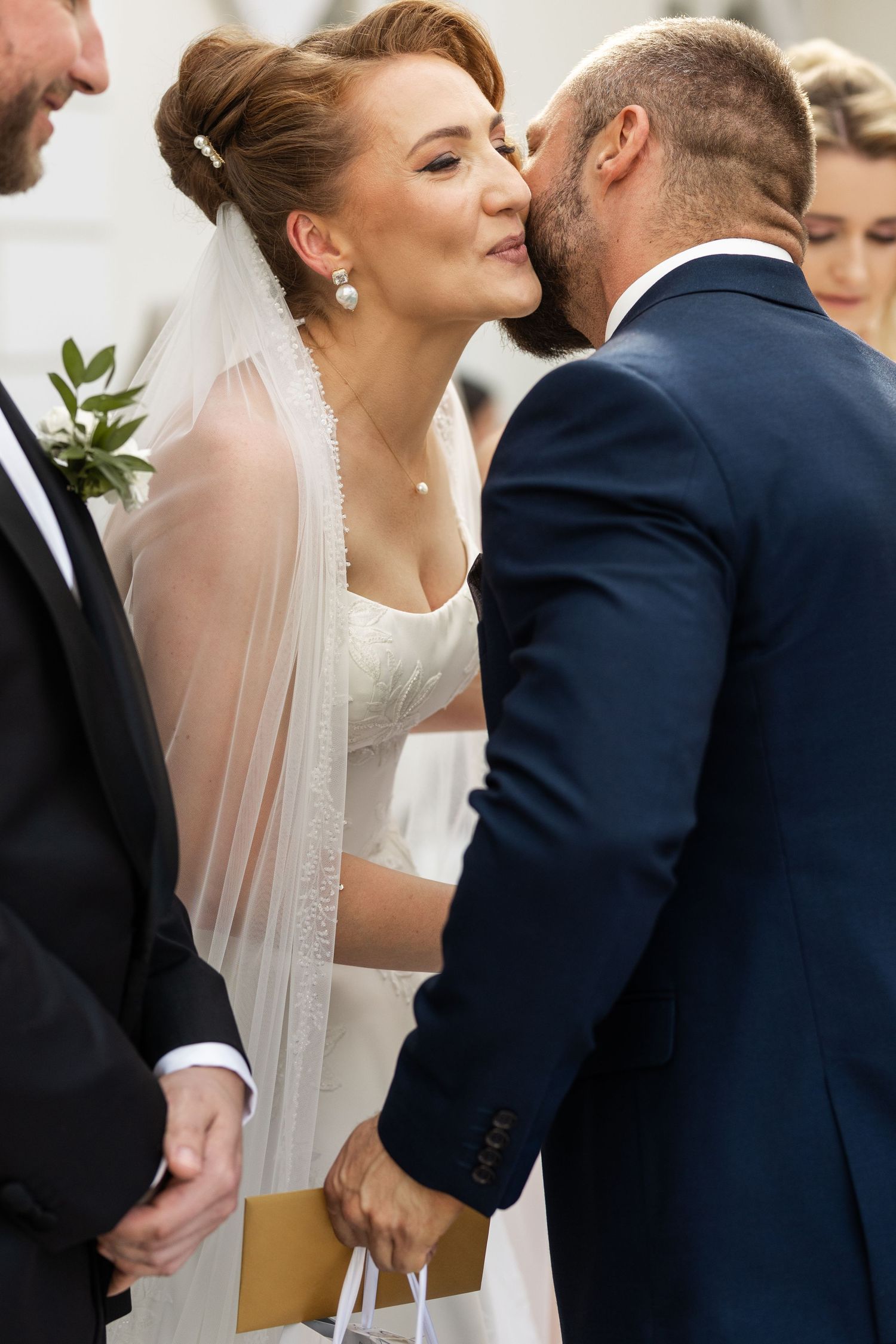 A bride in a white veil leans in for a kiss with her groom wearing a navy suit during their wedding ceremony.