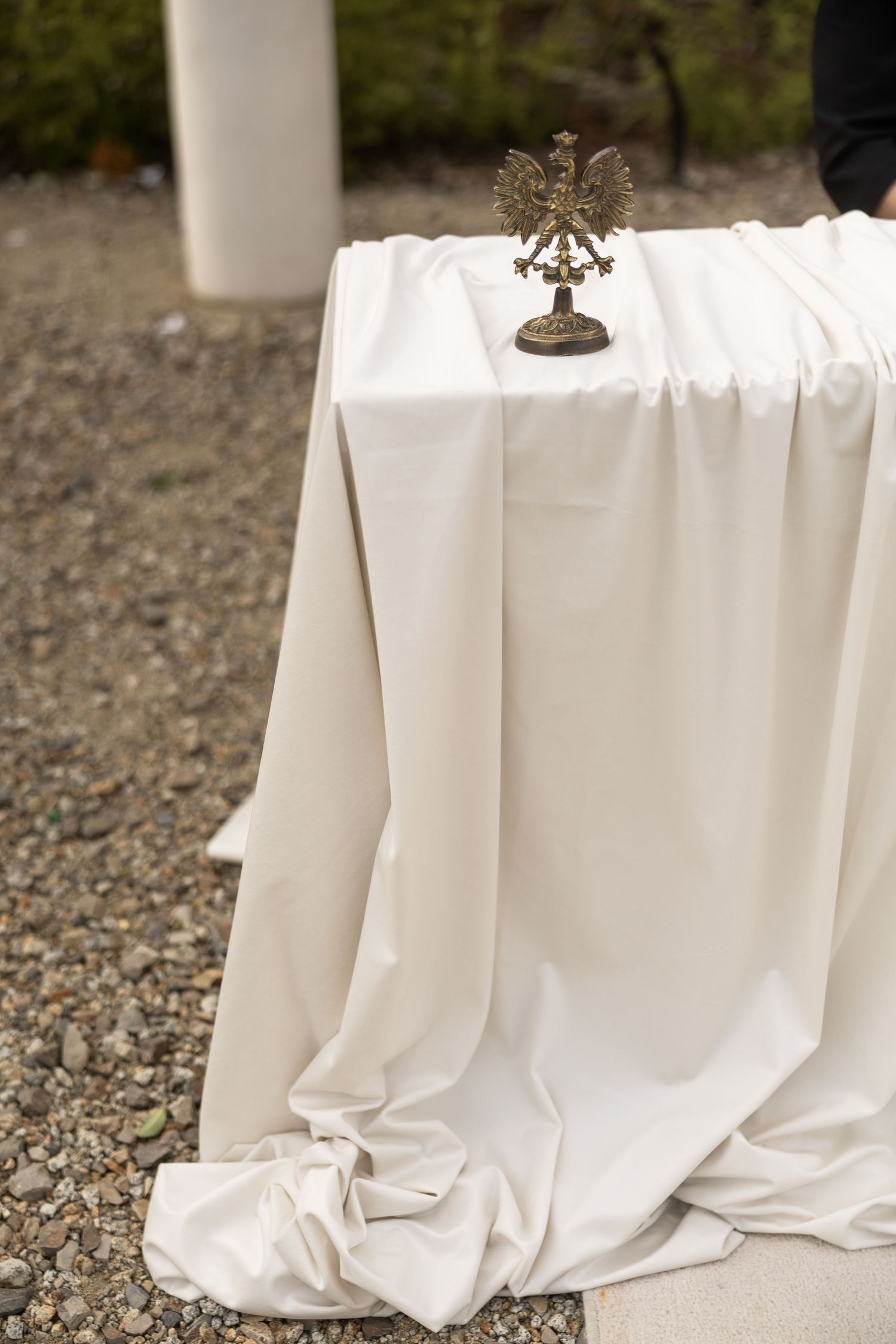 White draped table outdoors with ornate gold eagle figurine centerpiece on gravel ground.