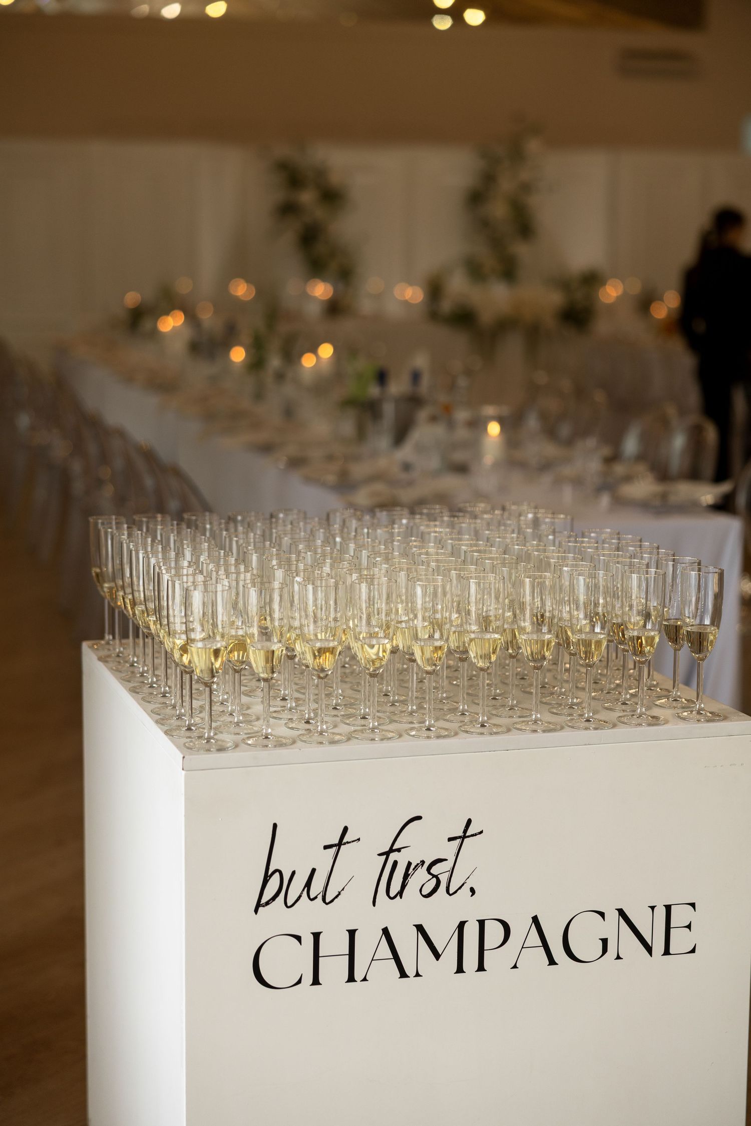 Champagne flutes arranged on a white pedestal with 'but first, CHAMPAGNE' text at an elegant wedding reception.