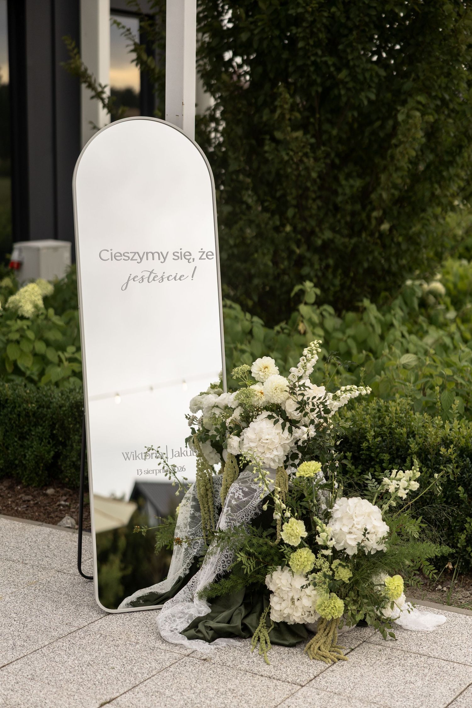 Elegant wedding welcome mirror with Polish text beside white floral arrangement on outdoor venue patio.
