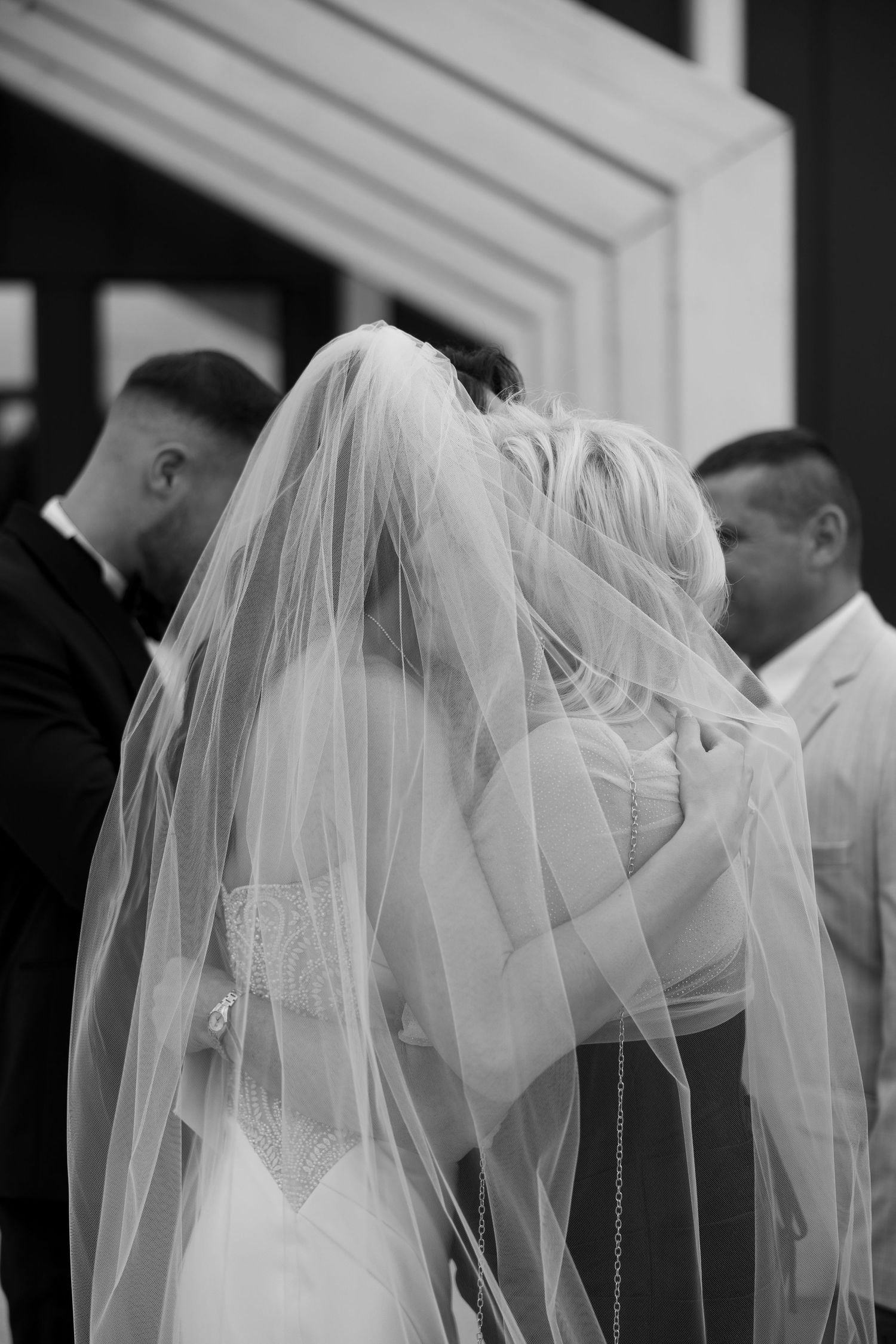 Black and white photo of a bride in a flowing veil embracing someone at a wedding ceremony outdoors.