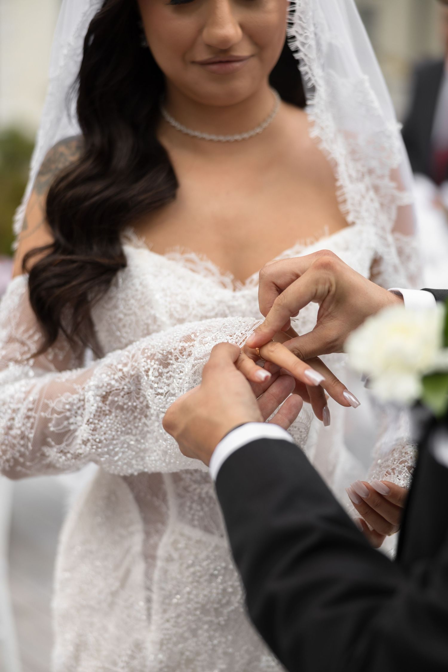 Bride in lace gown and veil receiving wedding ring from groom during outdoor ceremony.