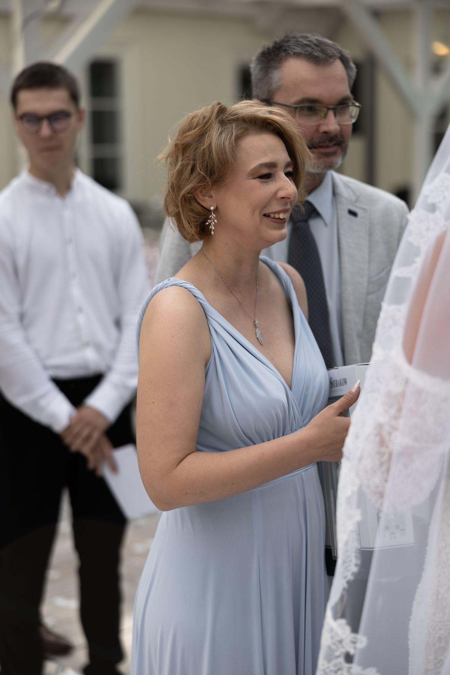 A smiling woman in a light blue bridesmaid dress adjusts a wedding veil at an outdoor ceremony.