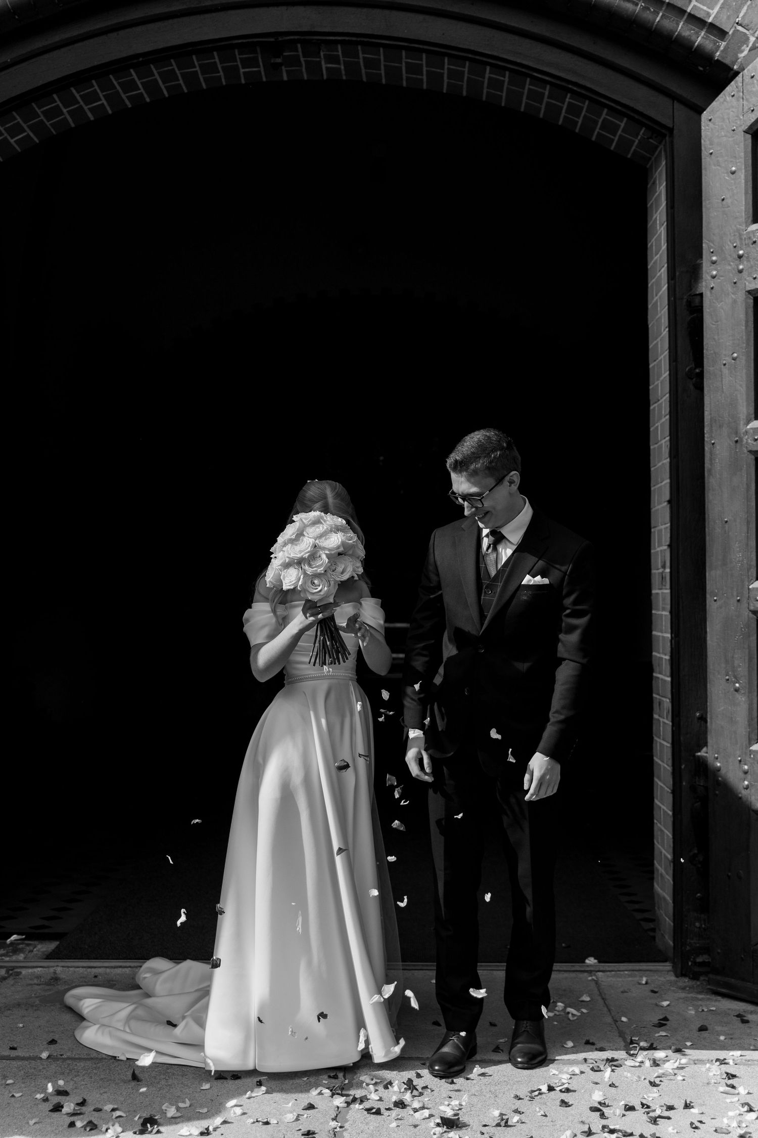 A black and white wedding scene showing a couple walking through scattered petals under an archway.