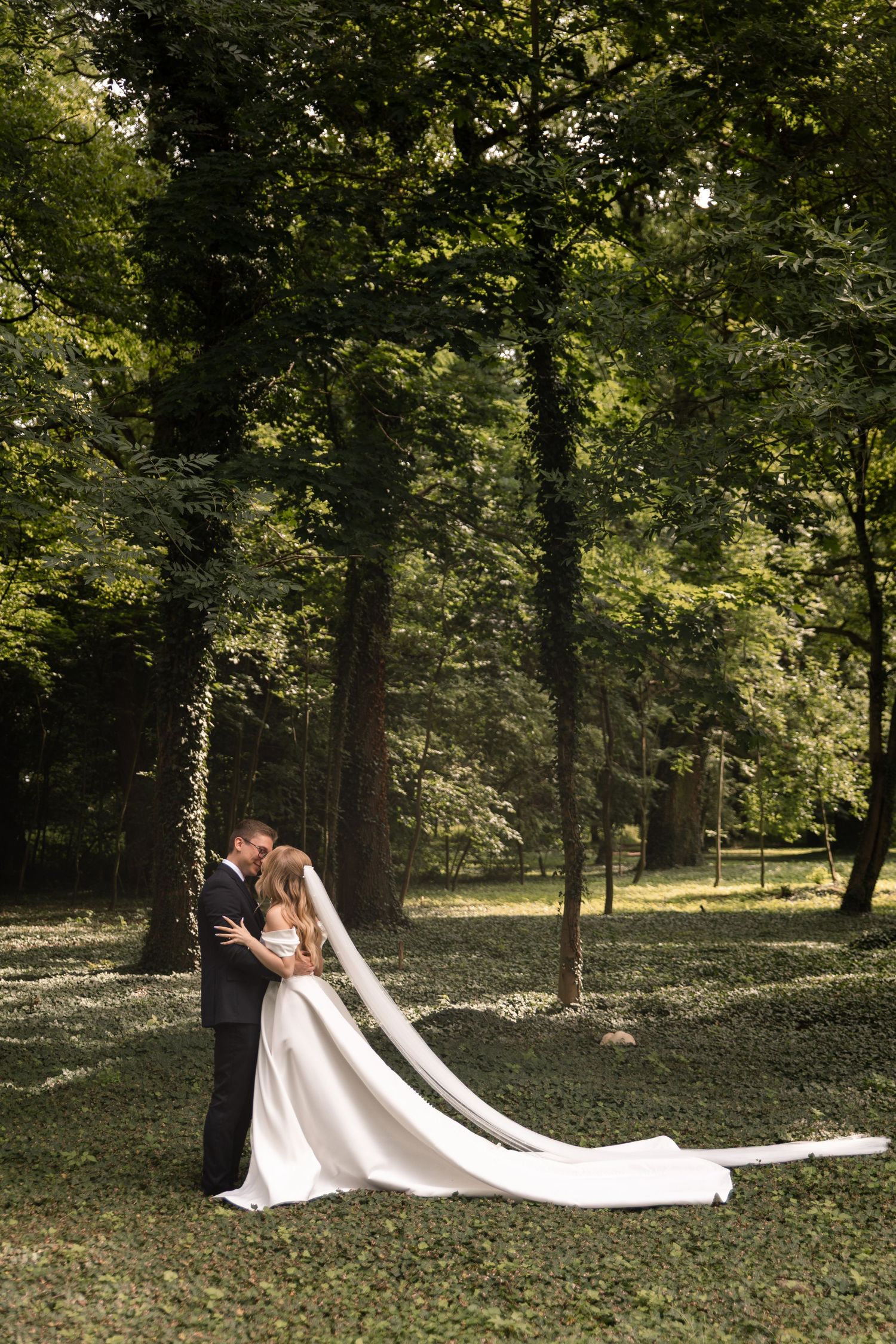 A romantic outdoor wedding photo series showing a couple embracing in a sunlit wooded setting with a flowing white dress train.