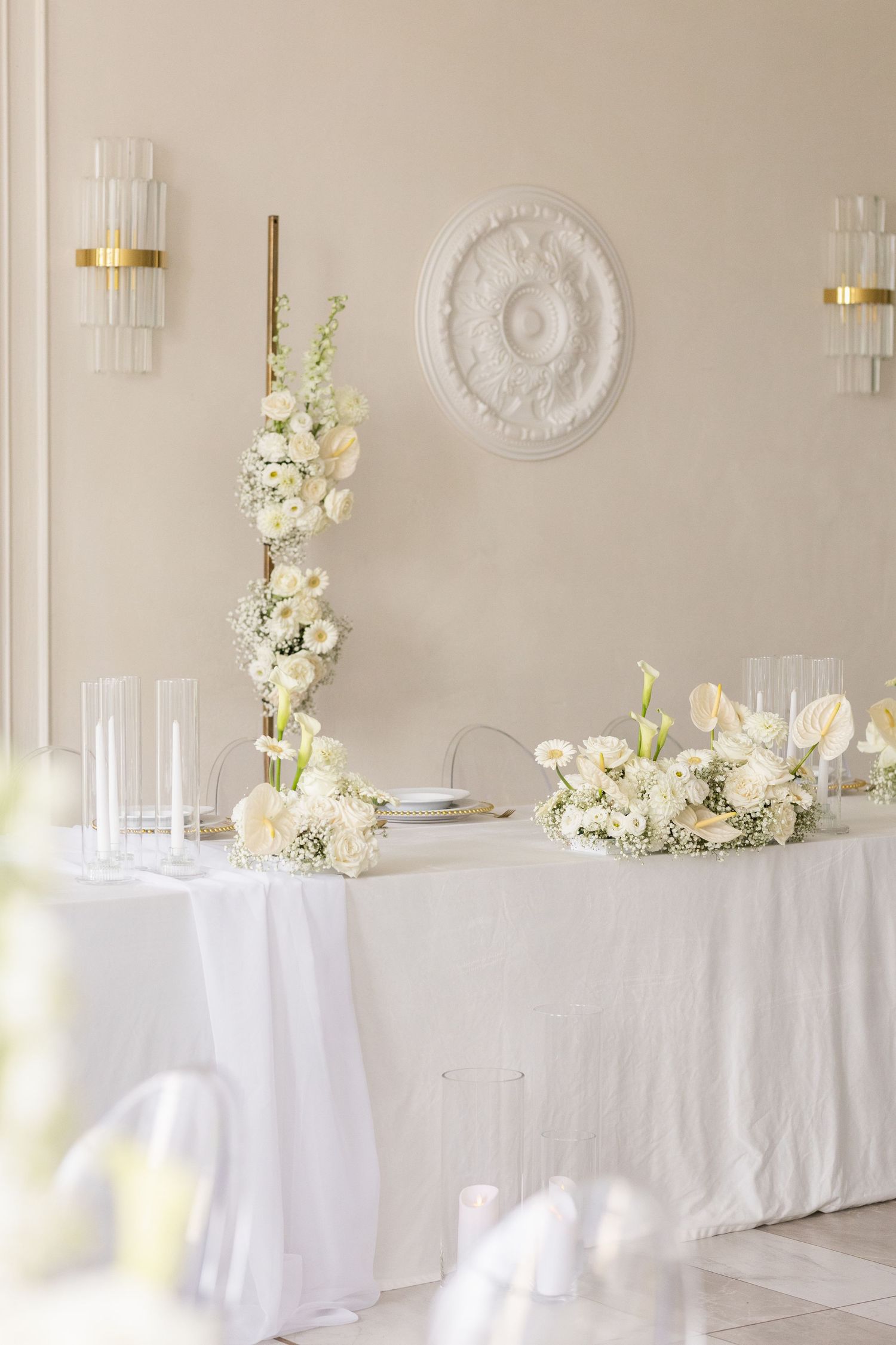 Elegant white wedding reception table with floral arrangements and wall sconces against beige wall with decorative medallion.