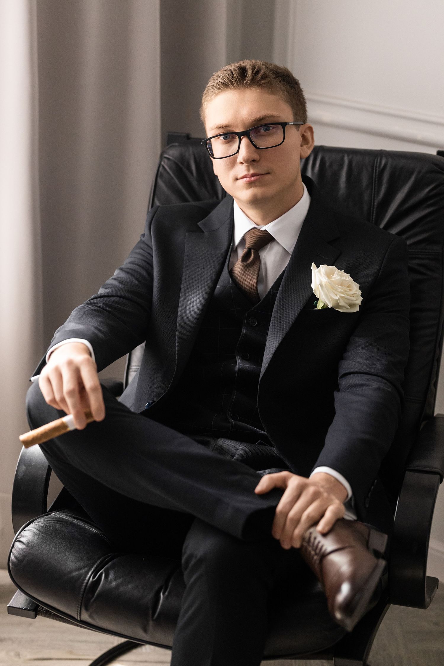 A person in a black suit and glasses sits in a leather chair holding a white rose boutonniere.