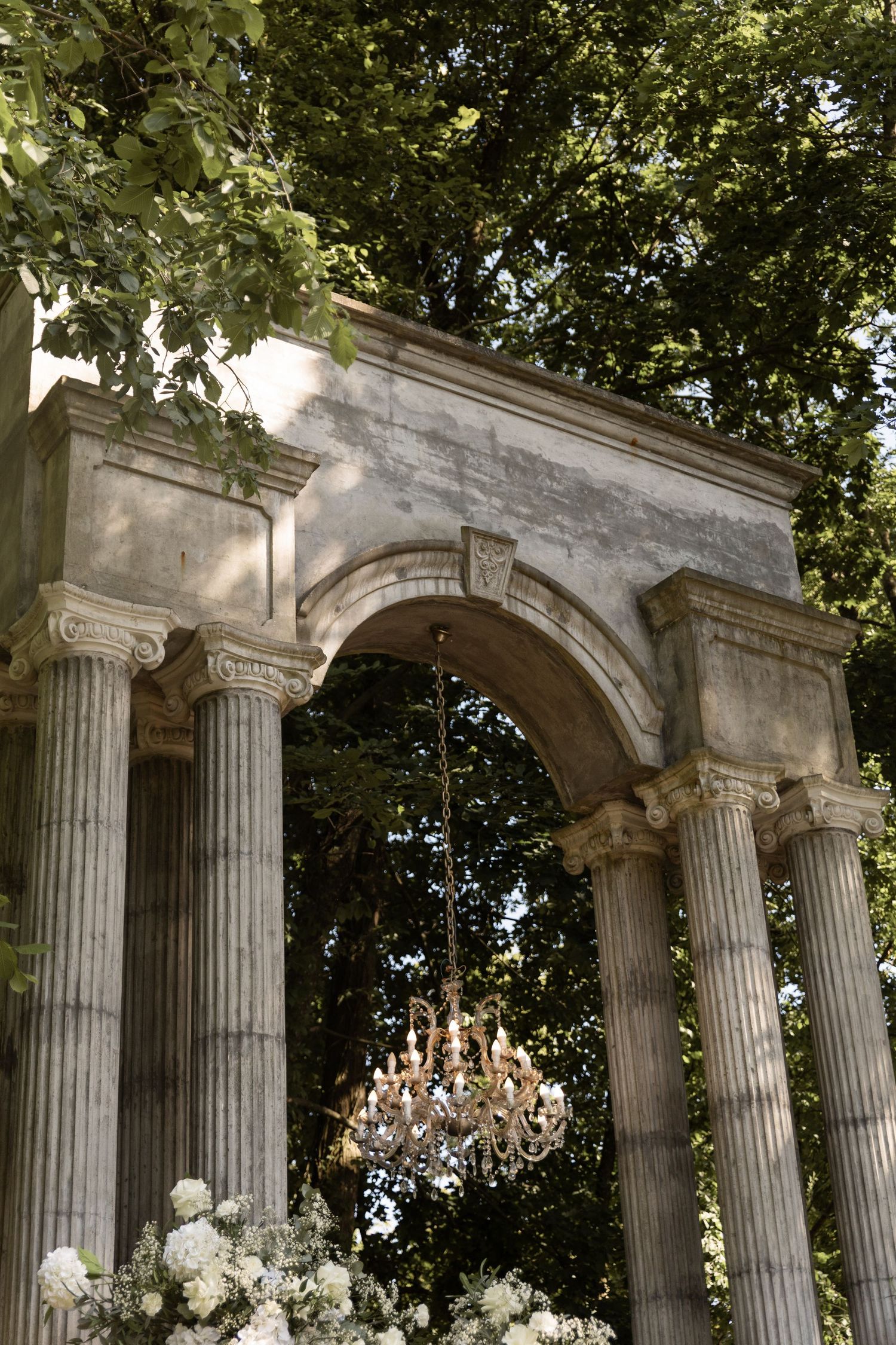 An elegant stone archway with Corinthian columns frames a hanging crystal chandelier in an outdoor garden setting.
