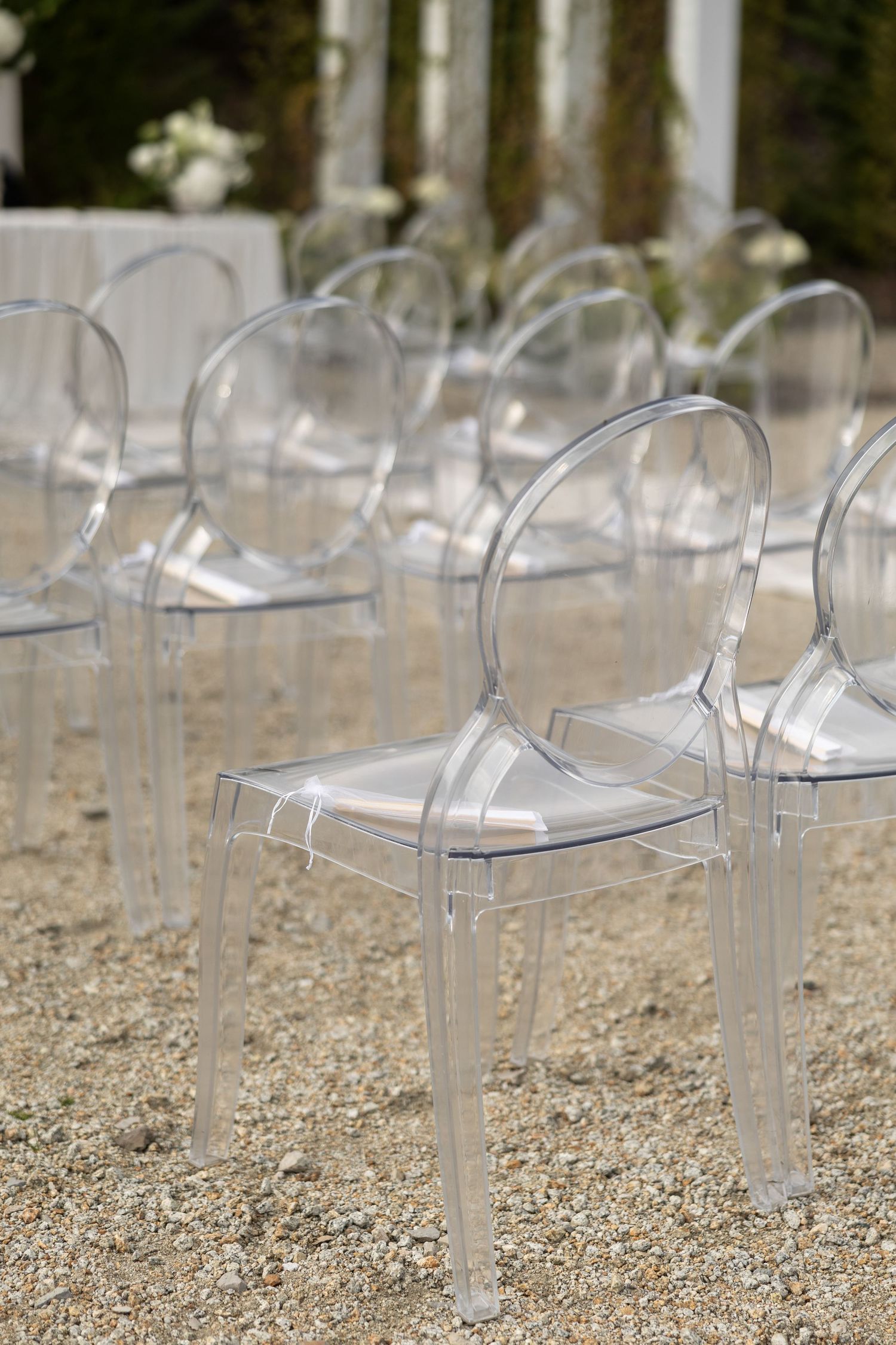 Elegant transparent ghost chairs arranged in rows on gravel for an outdoor wedding ceremony.
