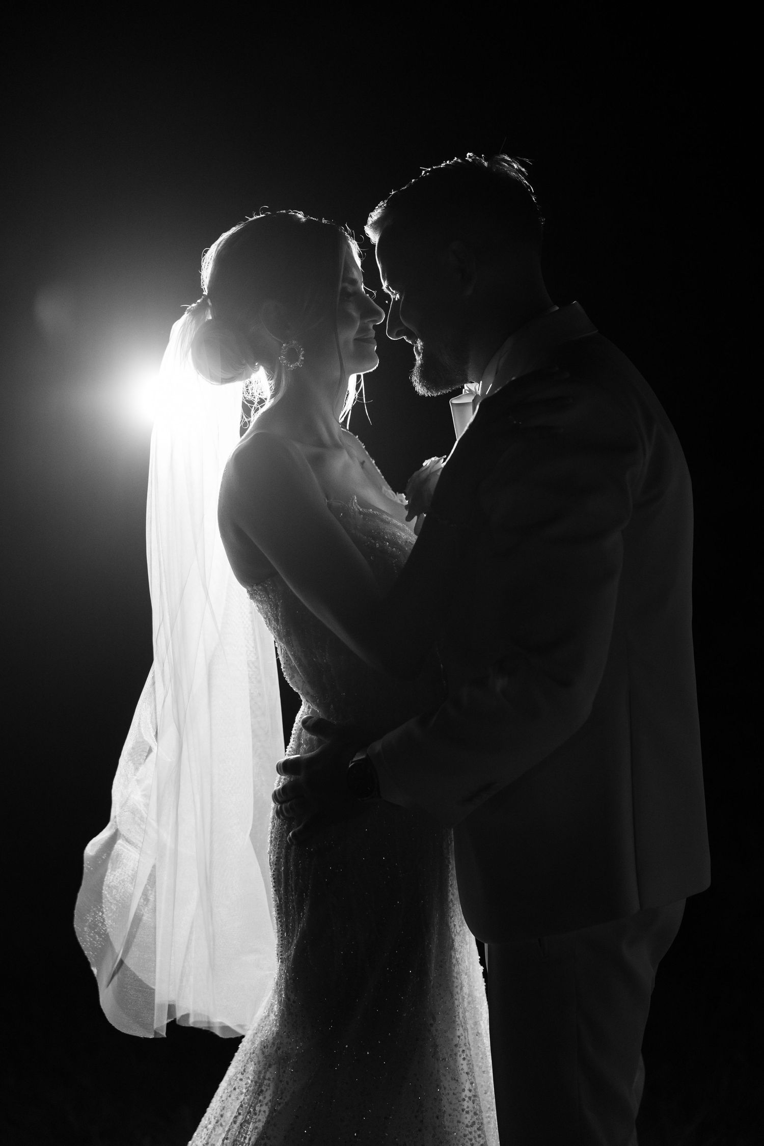 Black and white silhouette of bride and groom embracing with dramatic backlight on dark background.
