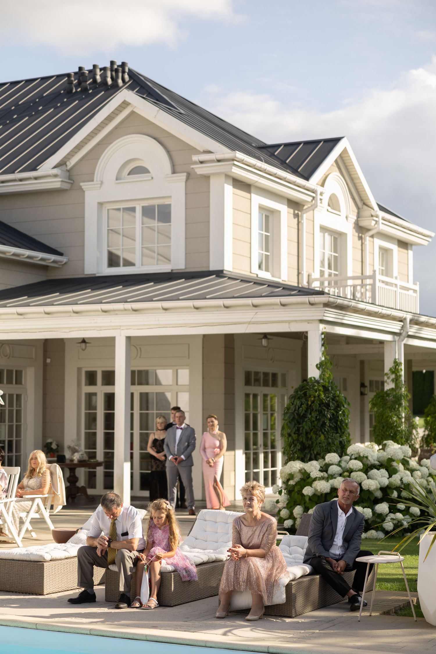 Elegant outdoor gathering at a luxury mansion with guests relaxing by a pool on a sunny day.