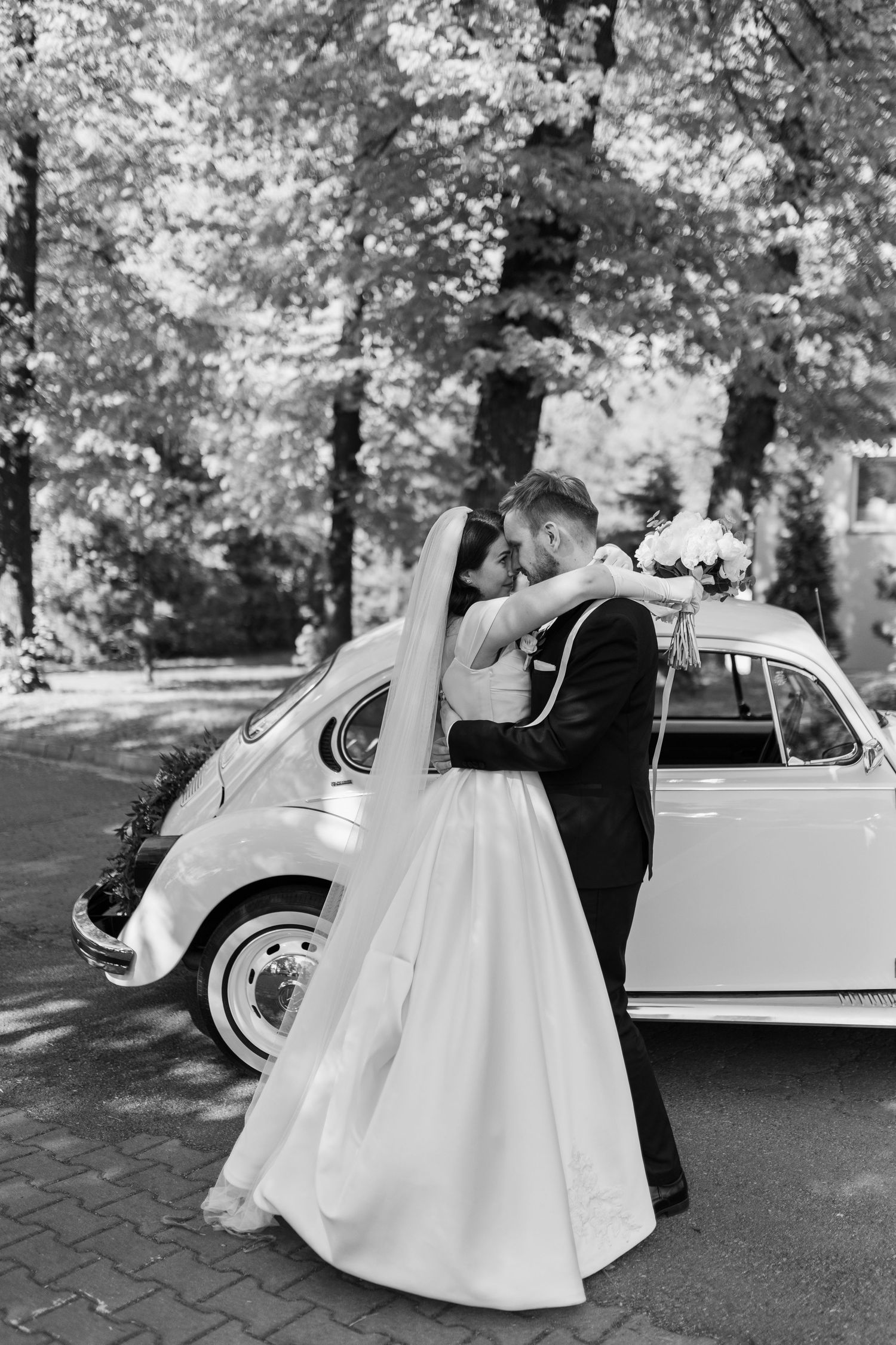 Black and white wedding photo with couple embracing beside vintage Volkswagen Beetle on tree-lined street.
