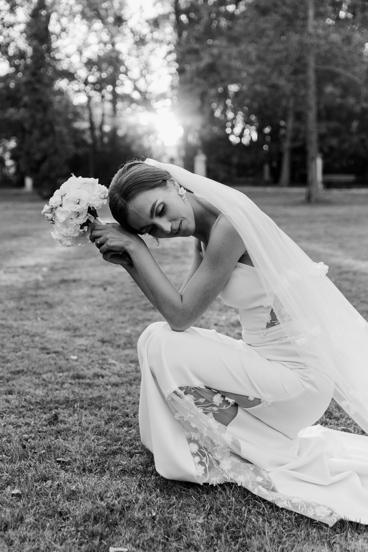 A bride in an elegant white dress kneels on grass holding a bouquet during a black and white outdoor photo session.
