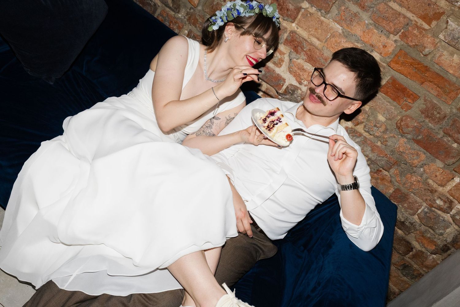 A newlywed couple in formal wedding attire playfully shares drinks against a rustic brick wall backdrop.