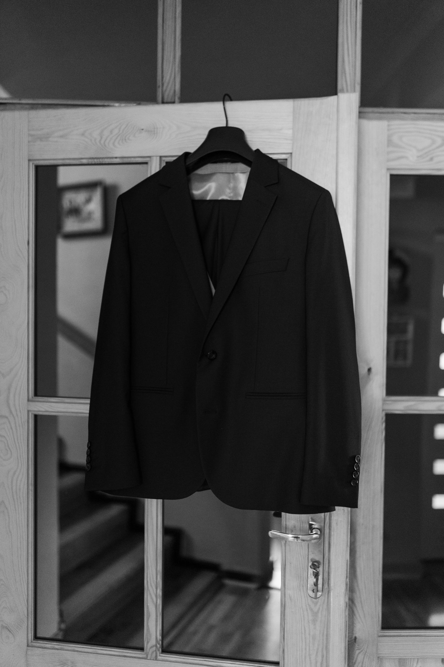 A black suit jacket hangs on a wooden hanger against a white paneled door in black and white photography.
