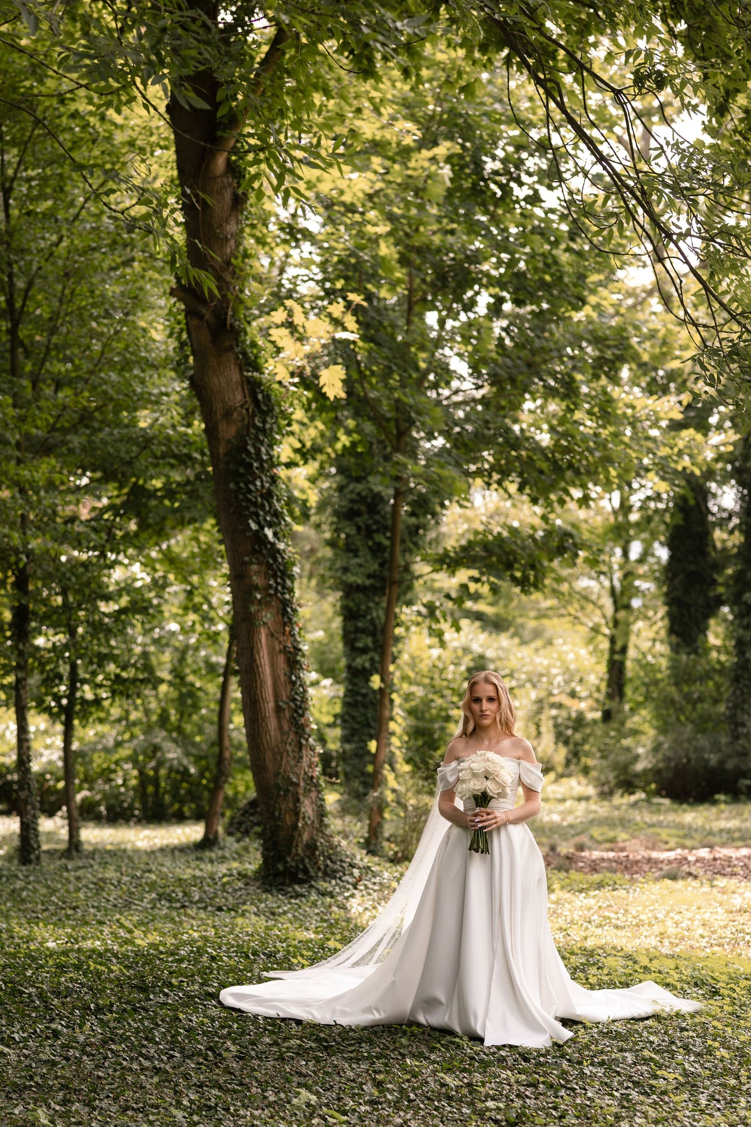 Two people in elegant white dresses embrace in a sunlit forest setting surrounded by lush greenery and trees.