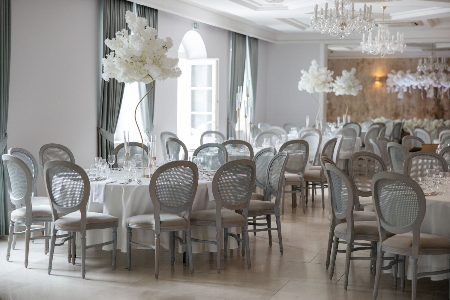 Elegant wedding reception hall with round tables, gray Louis chairs, white floral centerpieces, and crystal chandeliers.