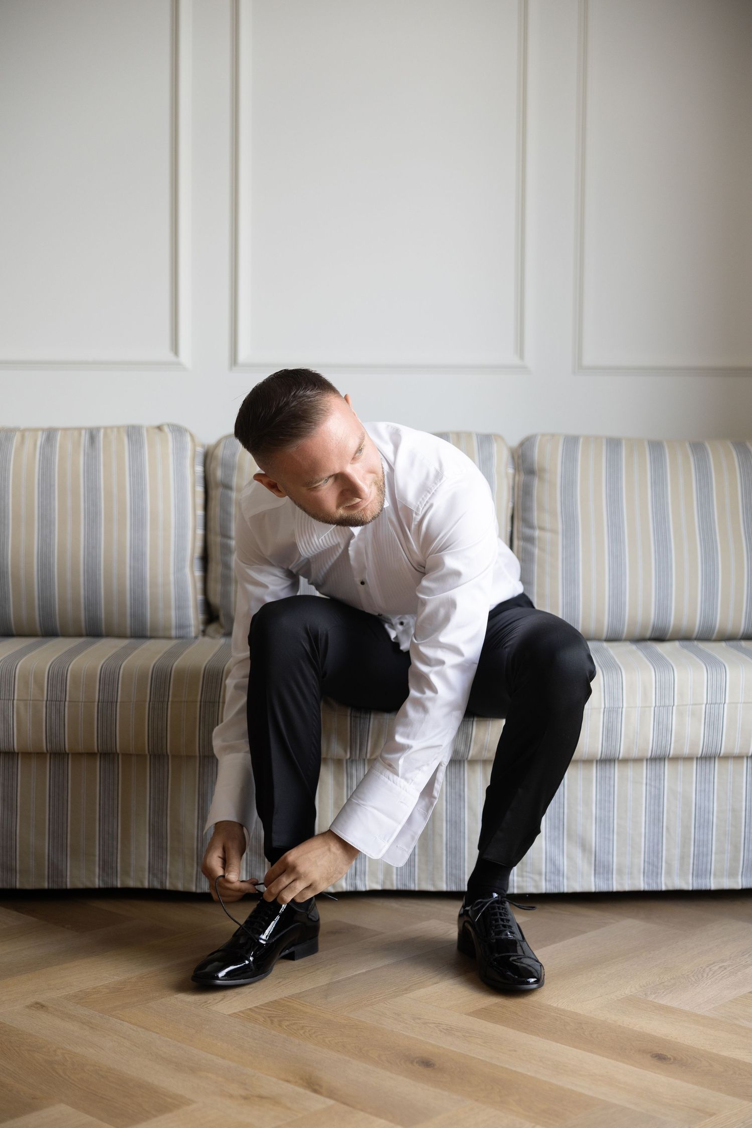 A well-dressed groom in white shirt and black trousers tying his shiny dress shoes while seated on a striped sofa.