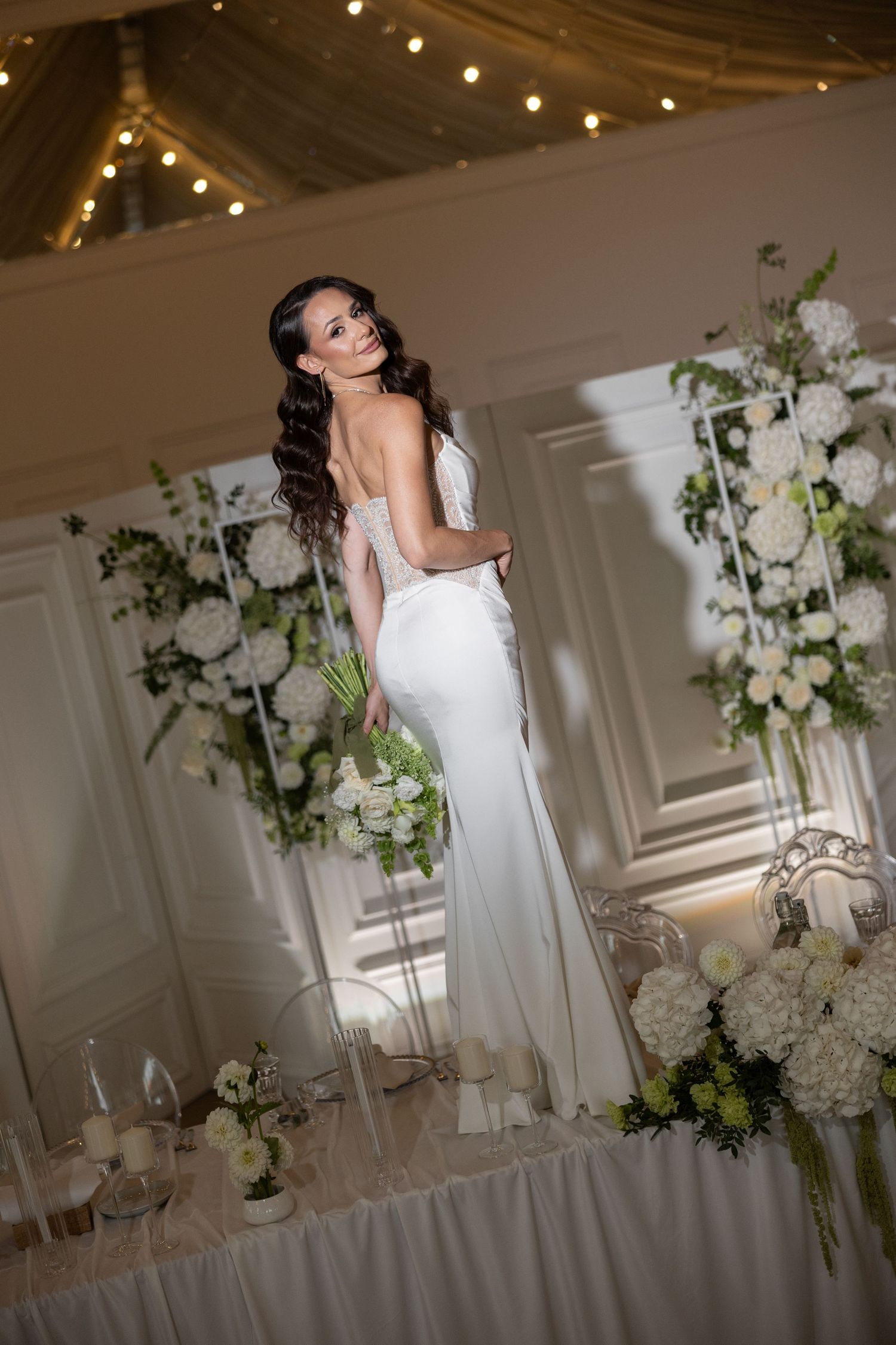 Elegant bride in a white backless gown holding a bouquet, surrounded by white floral arrangements in a decorated wedding venue.