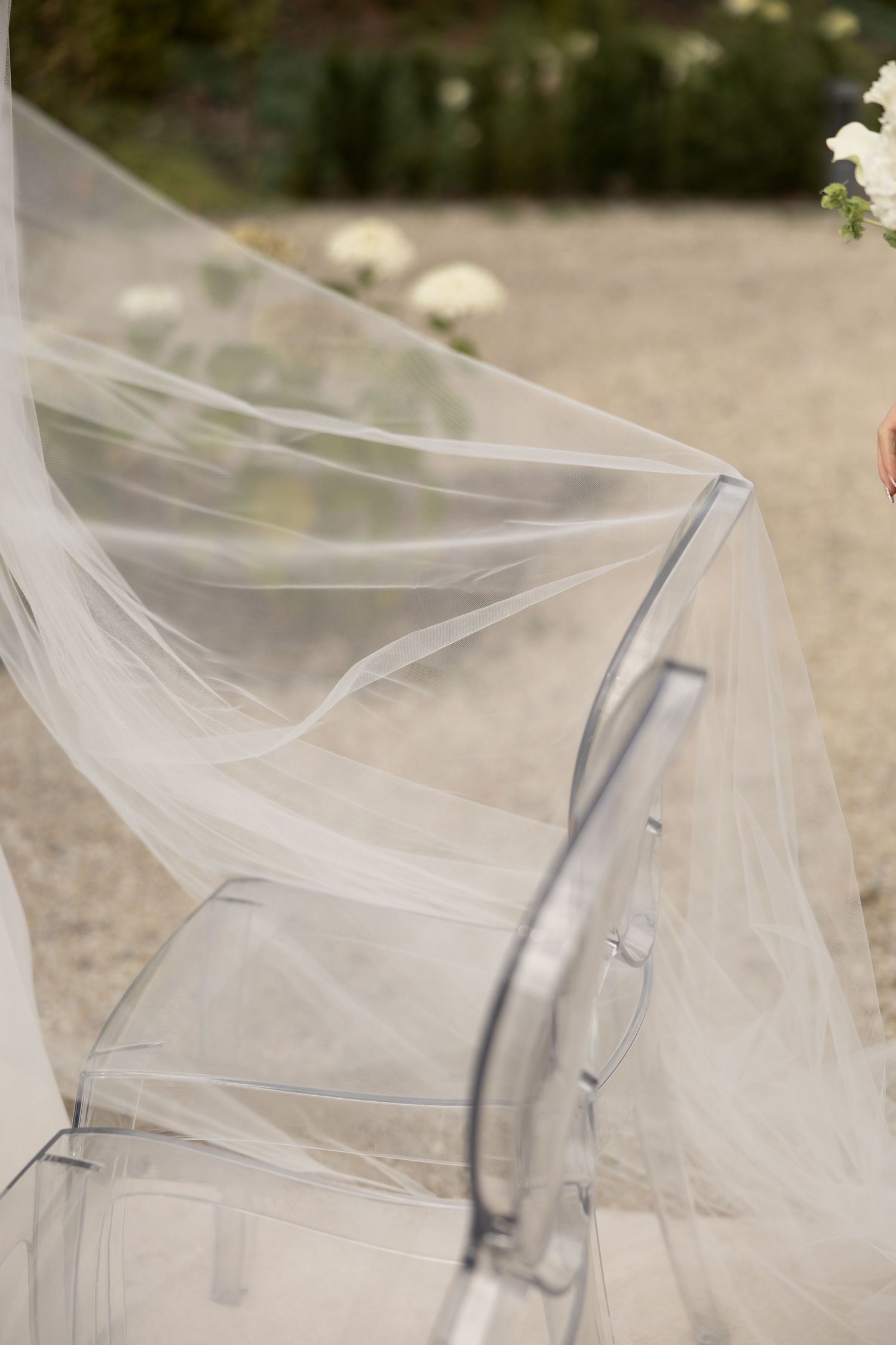 A flowing white bridal veil draped over a clear ghost chair at an outdoor wedding ceremony.