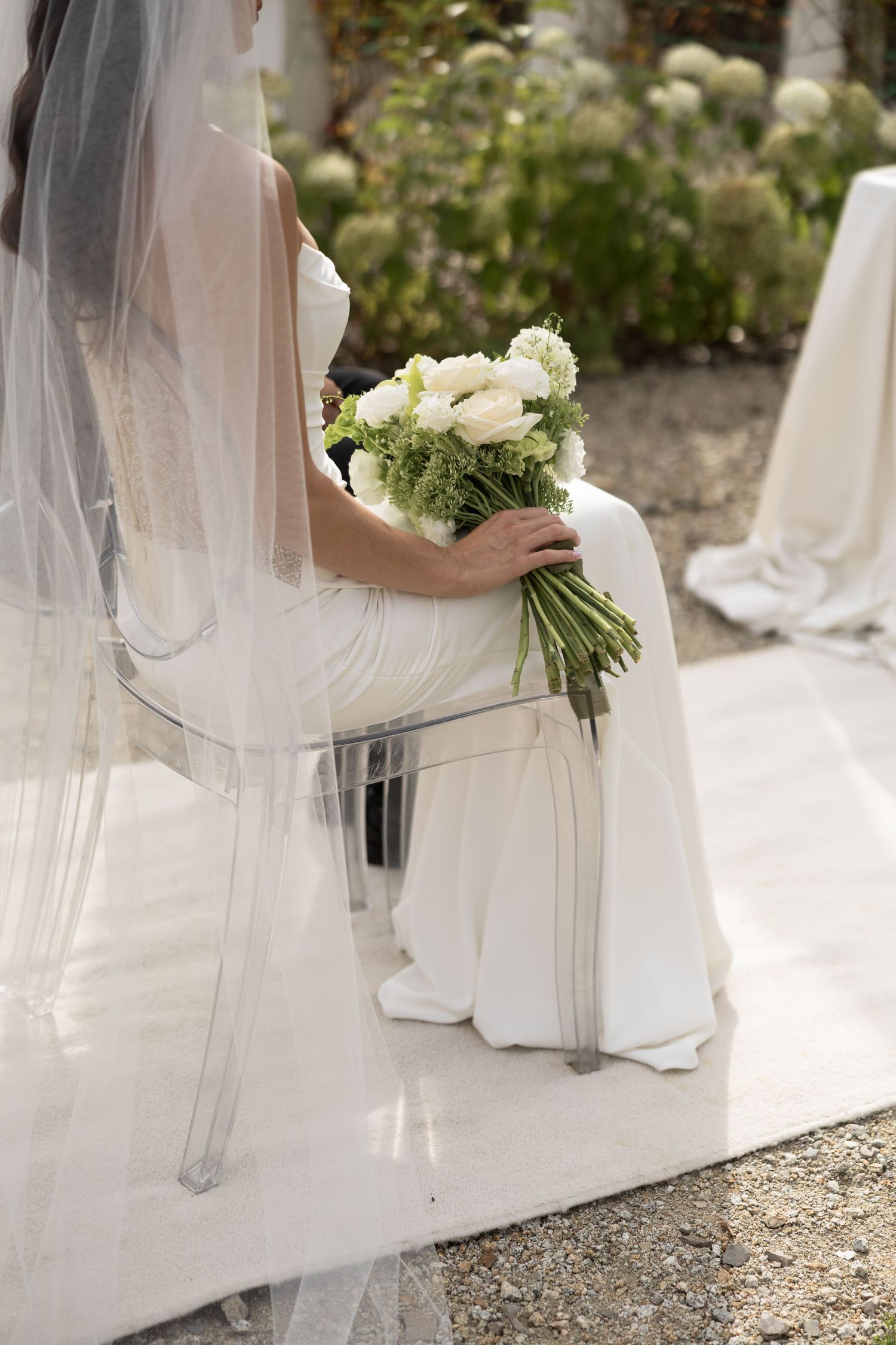 Bride in white gown and veil sits holding elegant white floral bouquet at outdoor wedding ceremony.