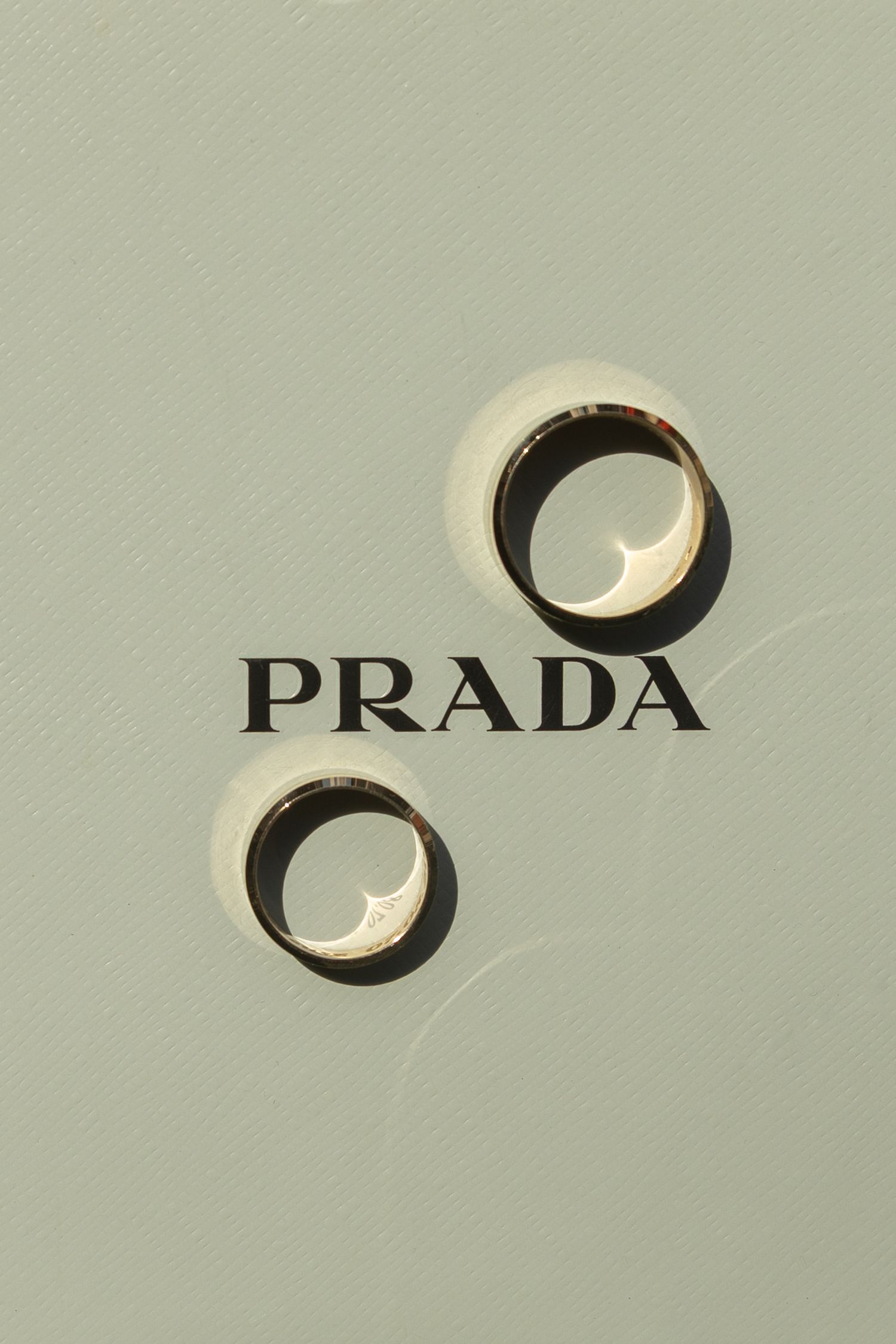 Two gold rings casting shadows on a sage green surface with the Prada logo printed in black between them.