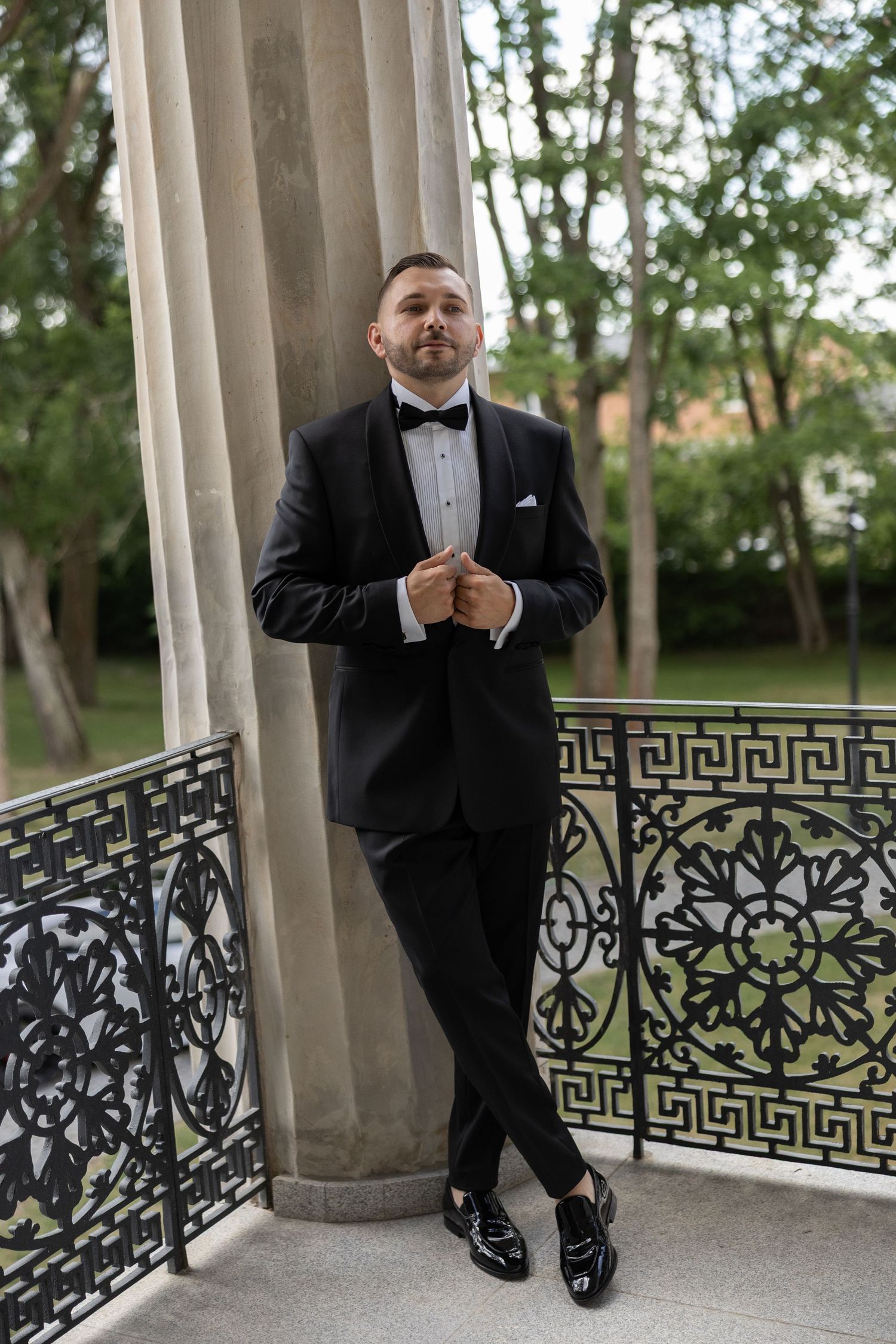 Elegant groom in black tuxedo and bow tie posing on ornate balcony with decorative iron railings.