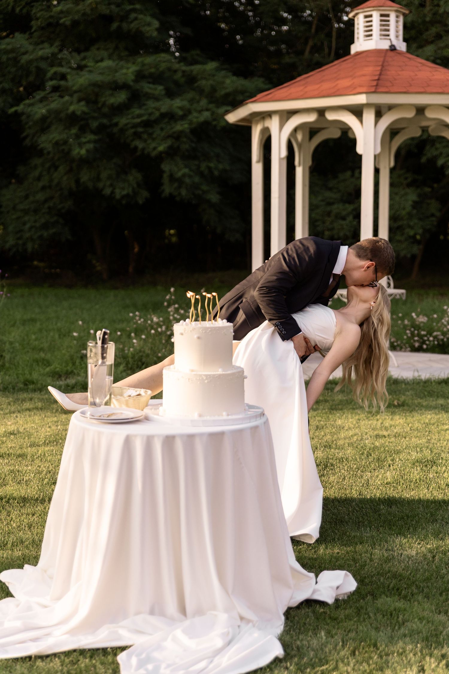 A couple shares a romantic kiss at a white-clothed table with a gazebo in the background during an outdoor event.