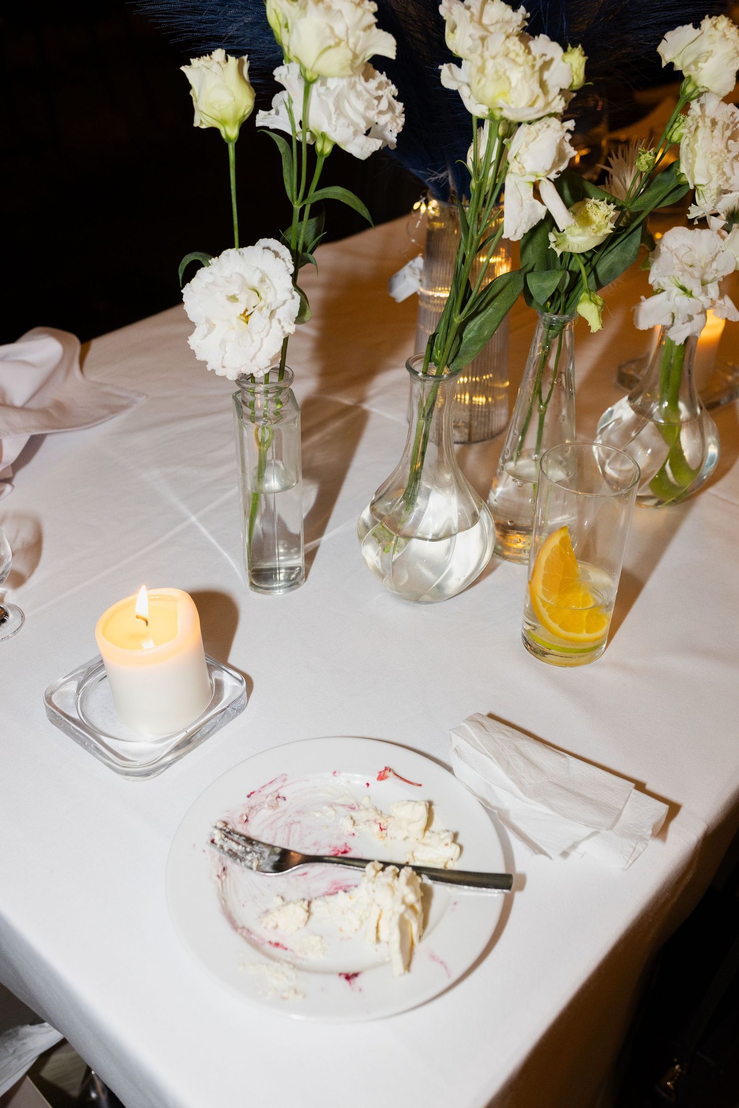 A table setting with white carnations in glass vases, a lit candle, and a partially eaten slice of cake on a white tablecloth.