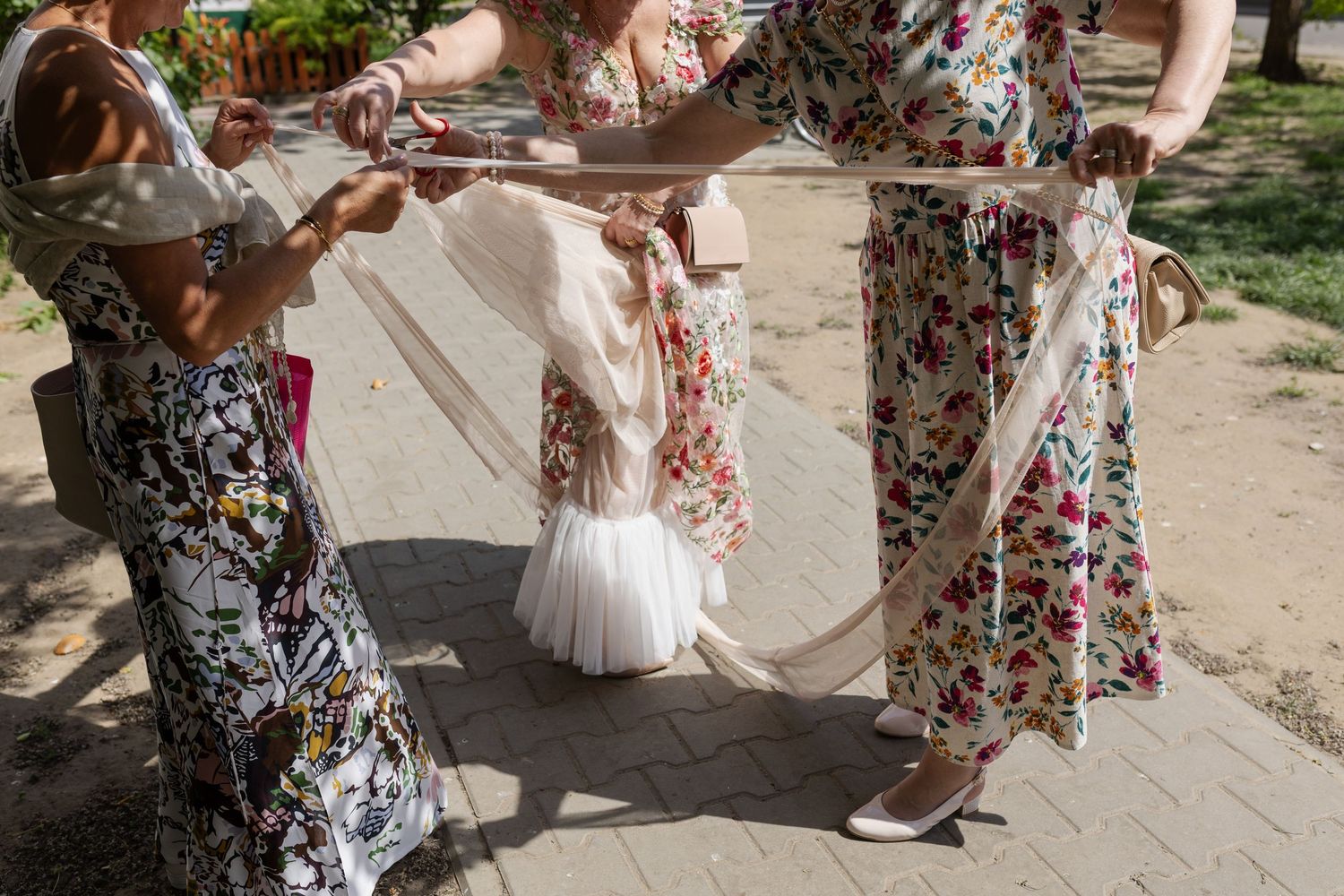 Guests in floral dresses assist with wedding dress preparations on a sunny outdoor walkway.
