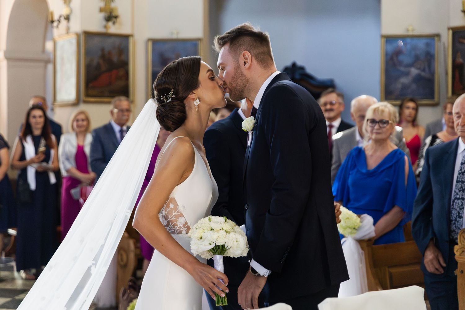A bride and groom share their first kiss during their wedding ceremony in a church with guests looking on.