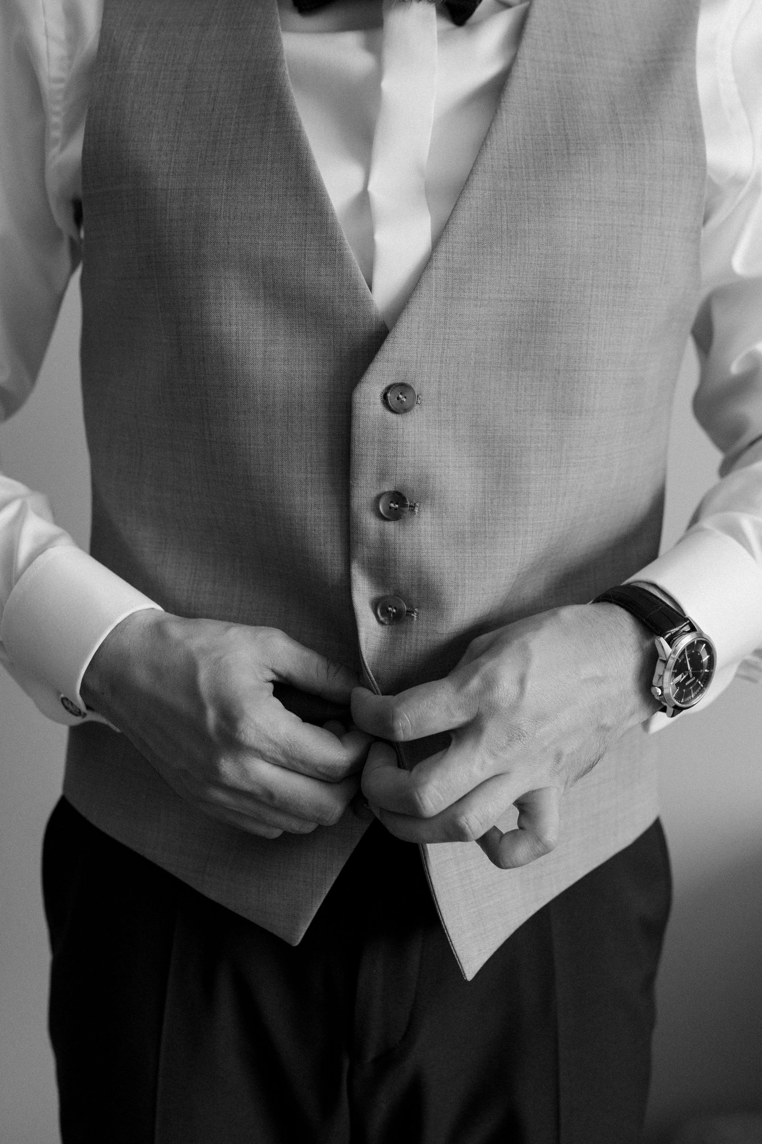 Black and white image of someone buttoning a formal gray vest with a wristwatch visible, showing elegant wedding attire details.