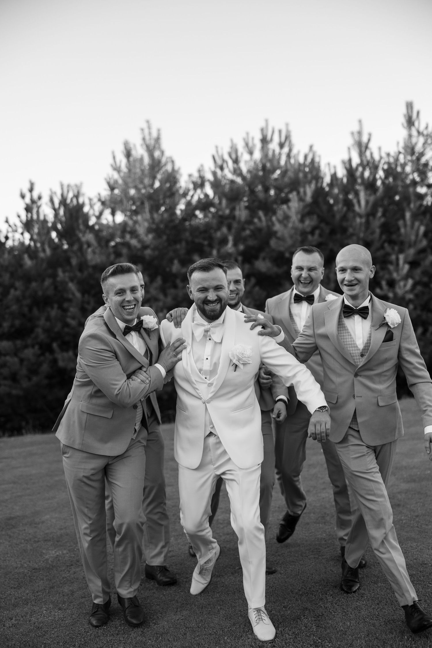 Black and white photo of a groom in white suit laughing with groomsmen in gray suits outdoors.