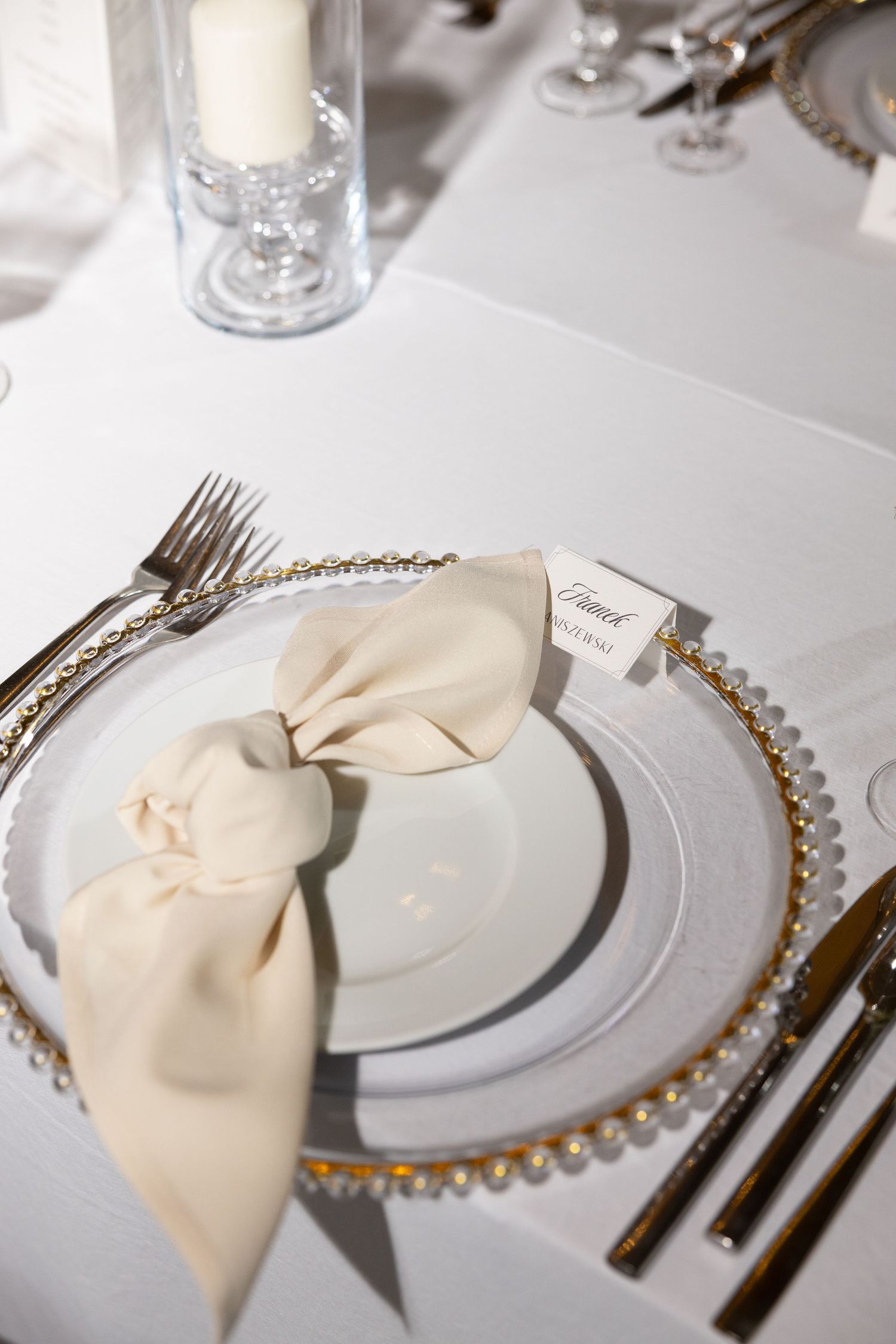 Elegant wedding table setting with gold-rimmed charger plate, cream linen napkin bow, place card, and crystal glassware.