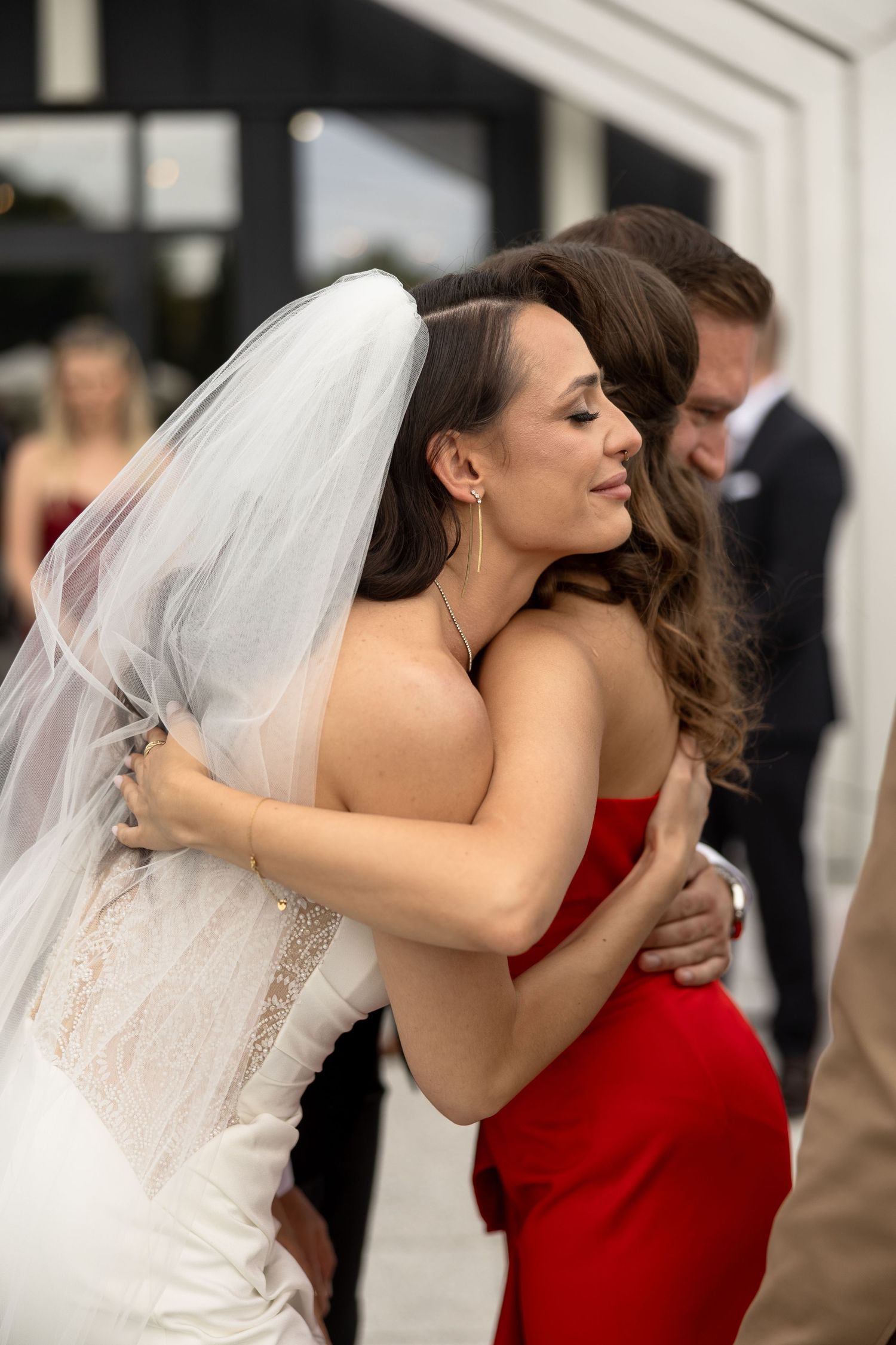 A bride in a white gown and veil shares a warm hug with a guest in a red dress at a wedding celebration.