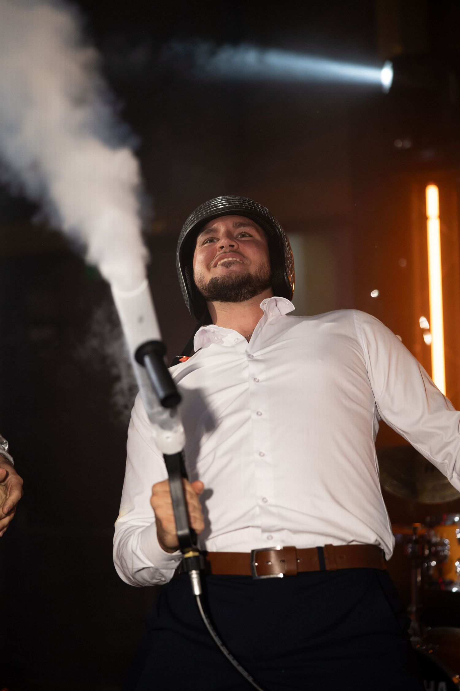Man in white shirt and helmet holds microphone on stage with smoke and dramatic lighting.