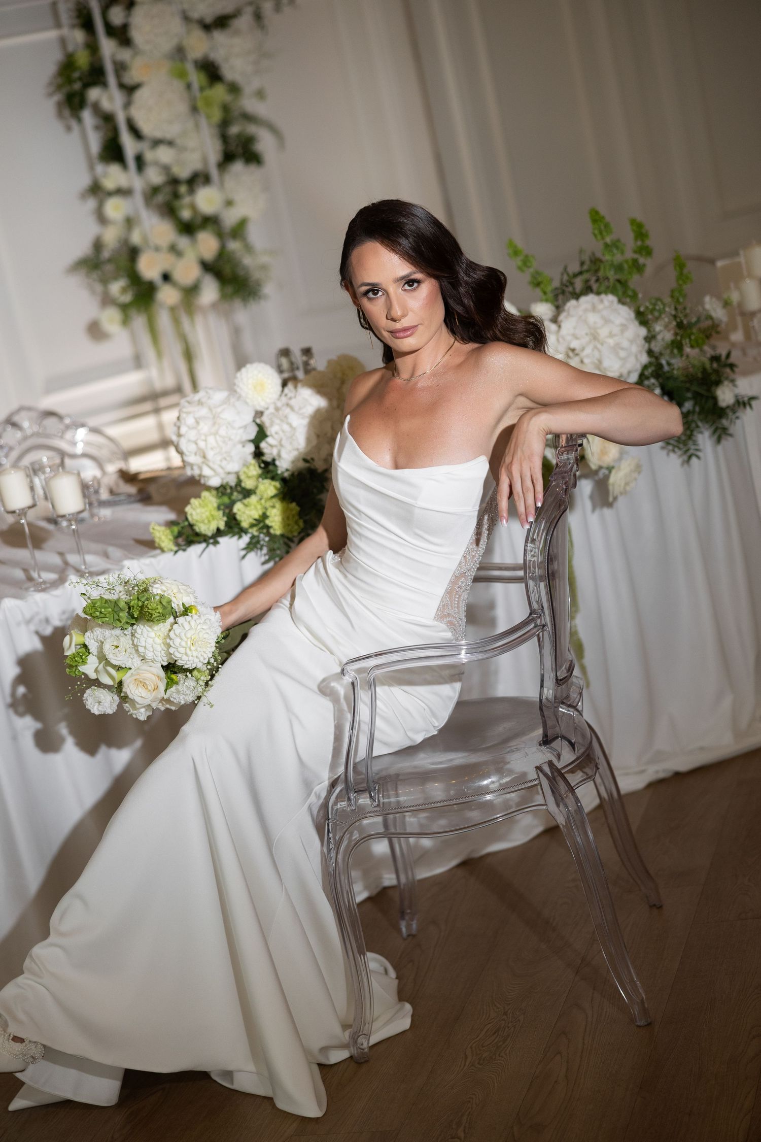 Elegant bride in strapless white gown seated on ghost chair holding bouquet at decorated wedding reception venue.