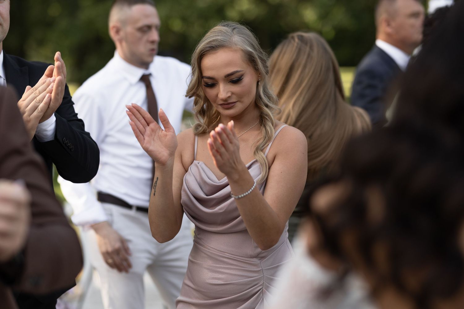 Elegant bridesmaid in silver dress clapping at outdoor wedding celebration surrounded by guests.