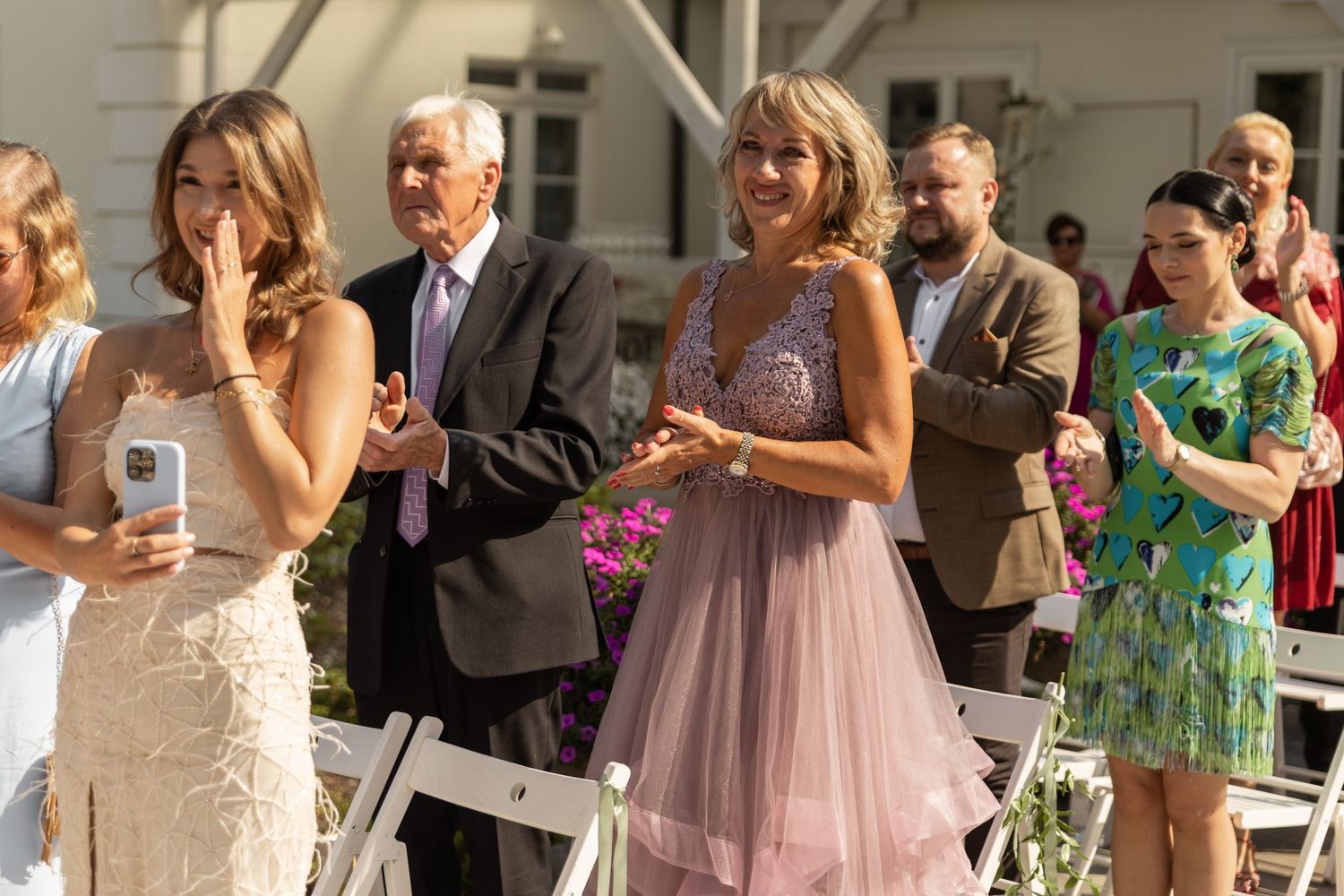 Wedding guests in formal attire stand in a line outdoors clapping and celebrating during a daytime ceremony.