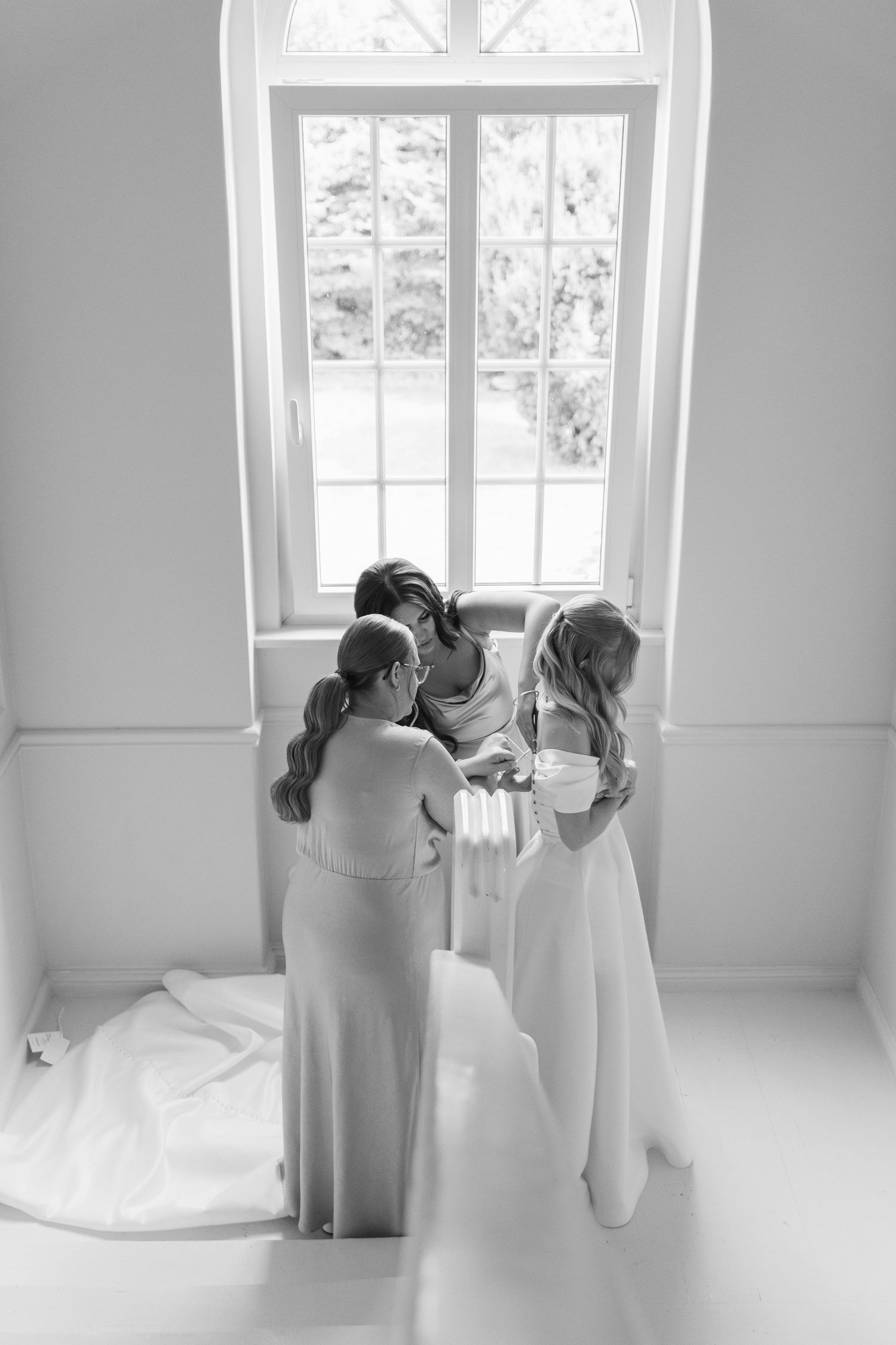 Black and white image showing bridesmaids helping with wedding preparations near bright windows in an elegant room.