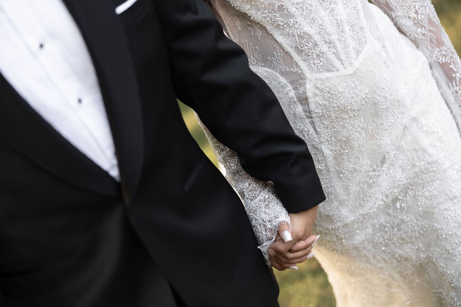 Close-up of bride and groom holding hands, showcasing wedding dress lace detail and black tuxedo suit.