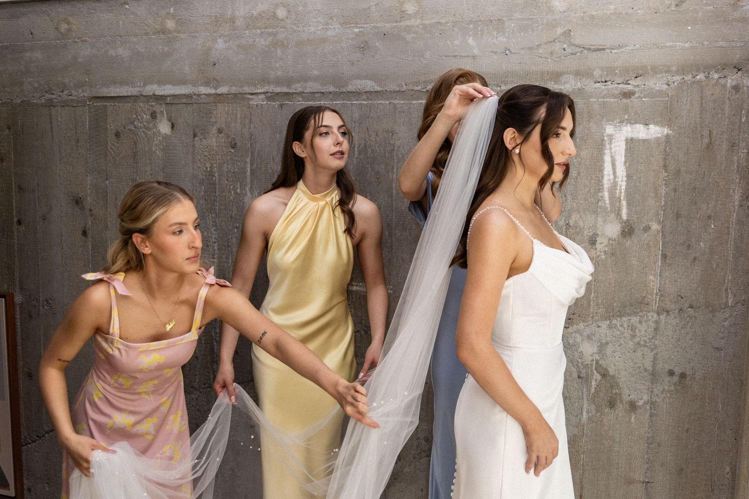 Bridesmaids in pastel dresses help adjust a bride's flowing veil against an industrial concrete wall backdrop.