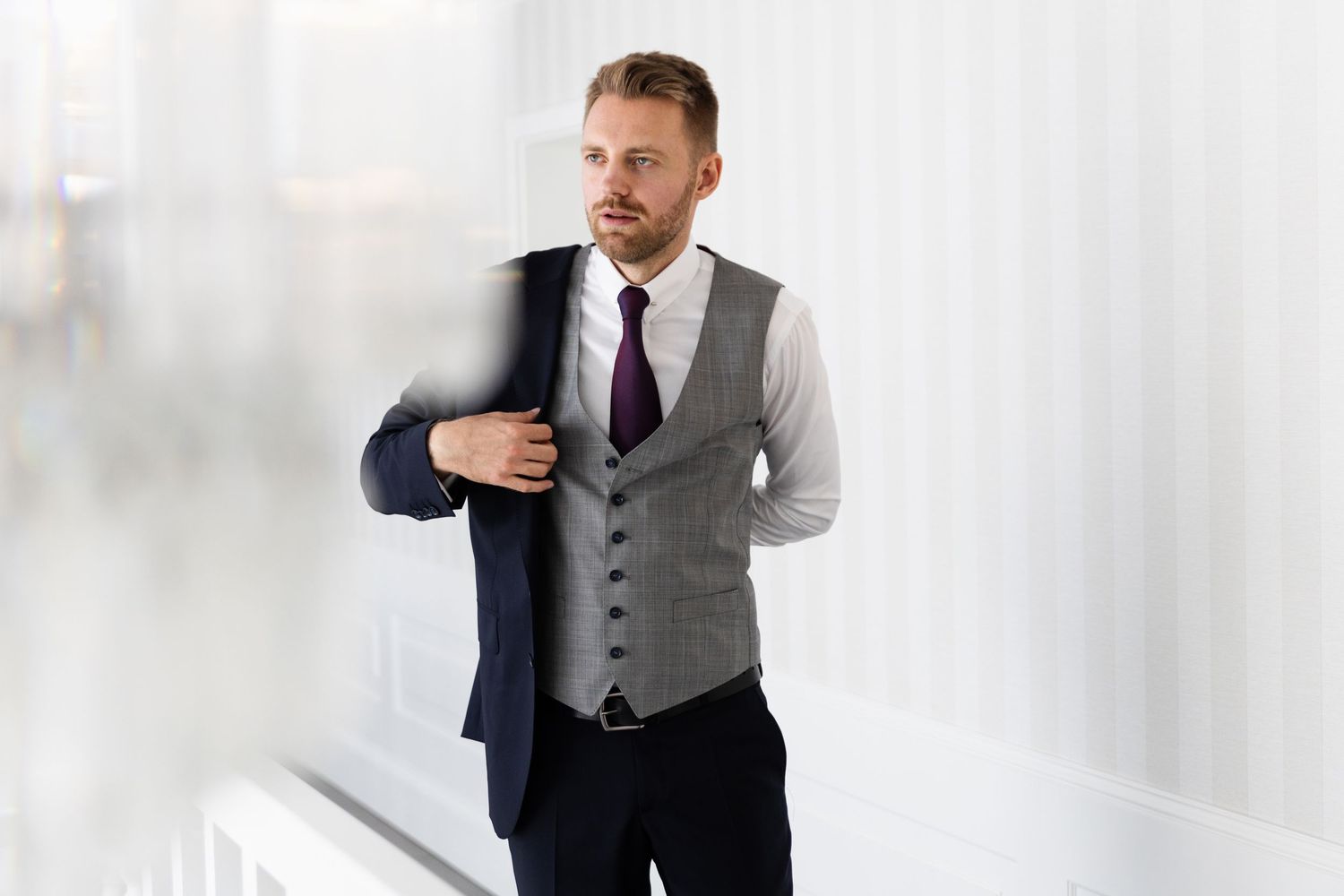 Businessman wearing grey waistcoat and navy blazer adjusts his jacket while standing in bright white interior.