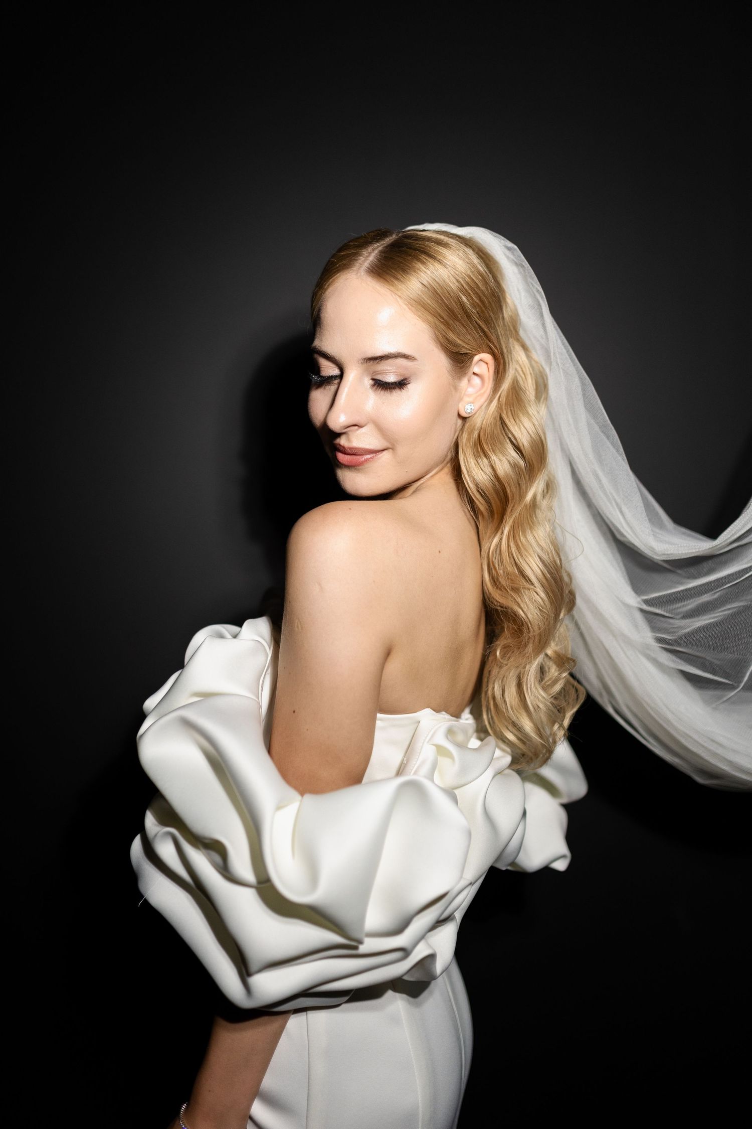 Bride in white dress with flowing veil poses against dark background.