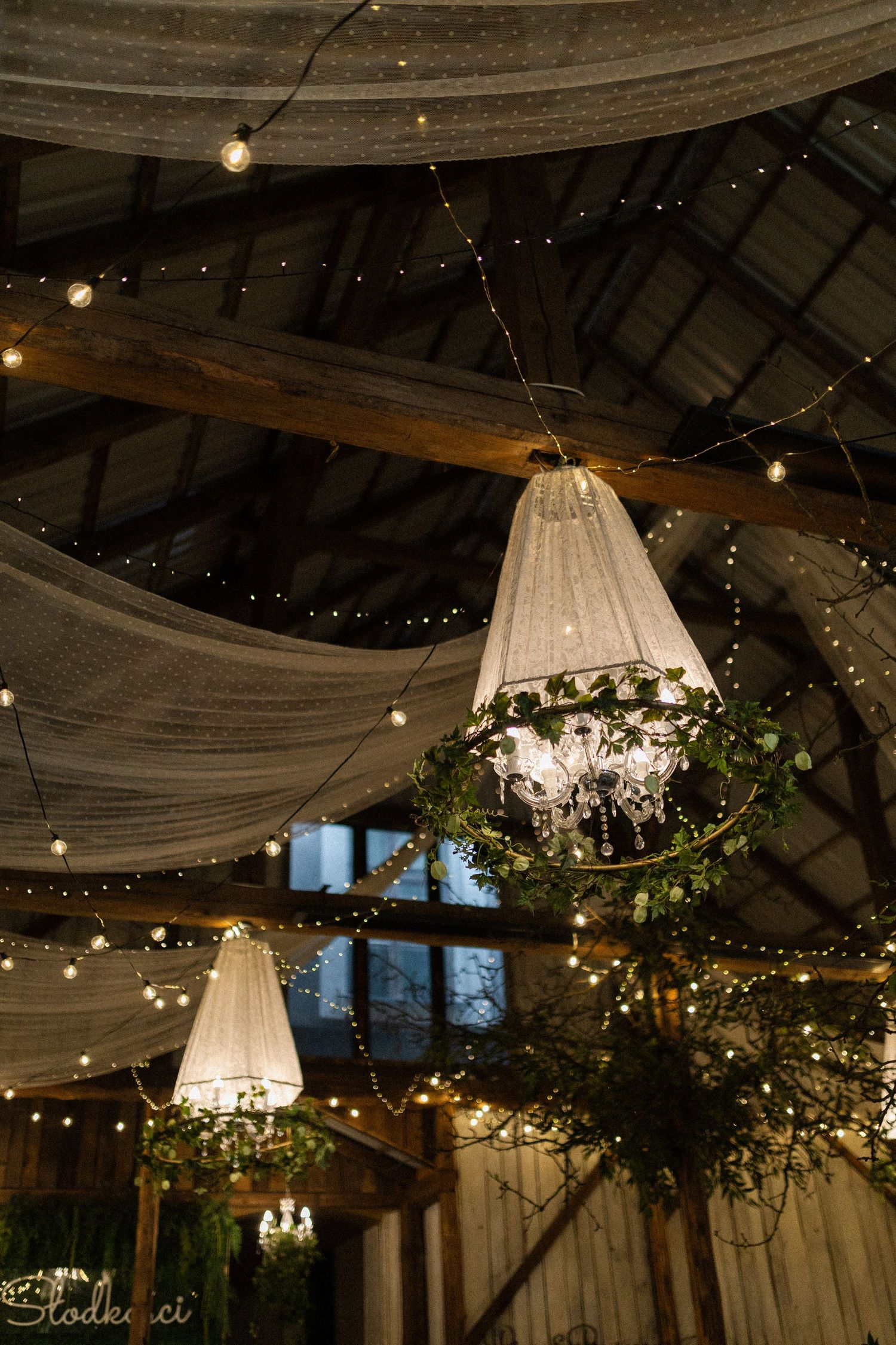 Elegant wedding venue interior with hanging light fixtures, draped fabric, and twinkling string lights adorning a rustic ceiling.