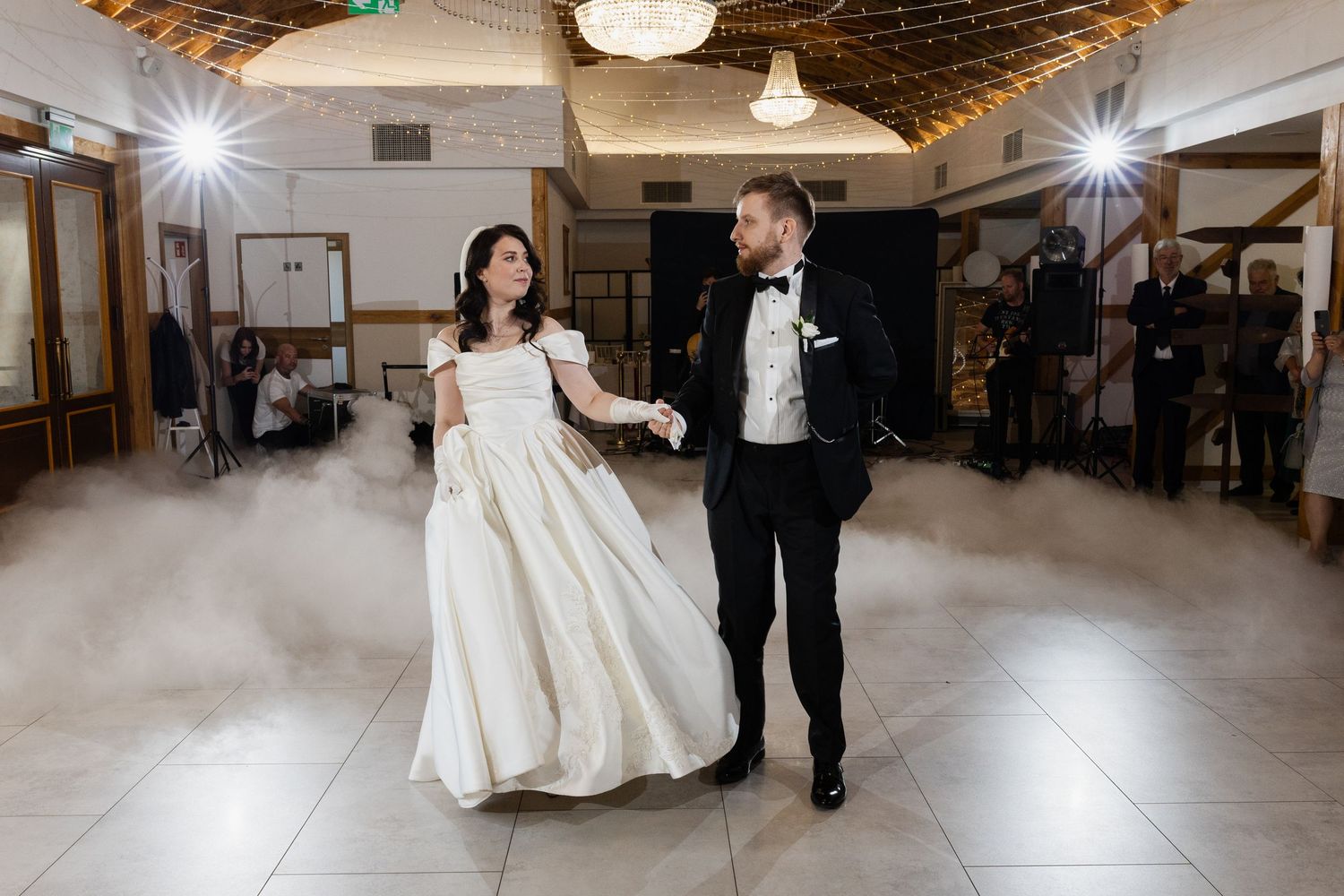 A newlywed couple dancing their first dance on a misty dance floor illuminated by overhead lights in a reception hall.