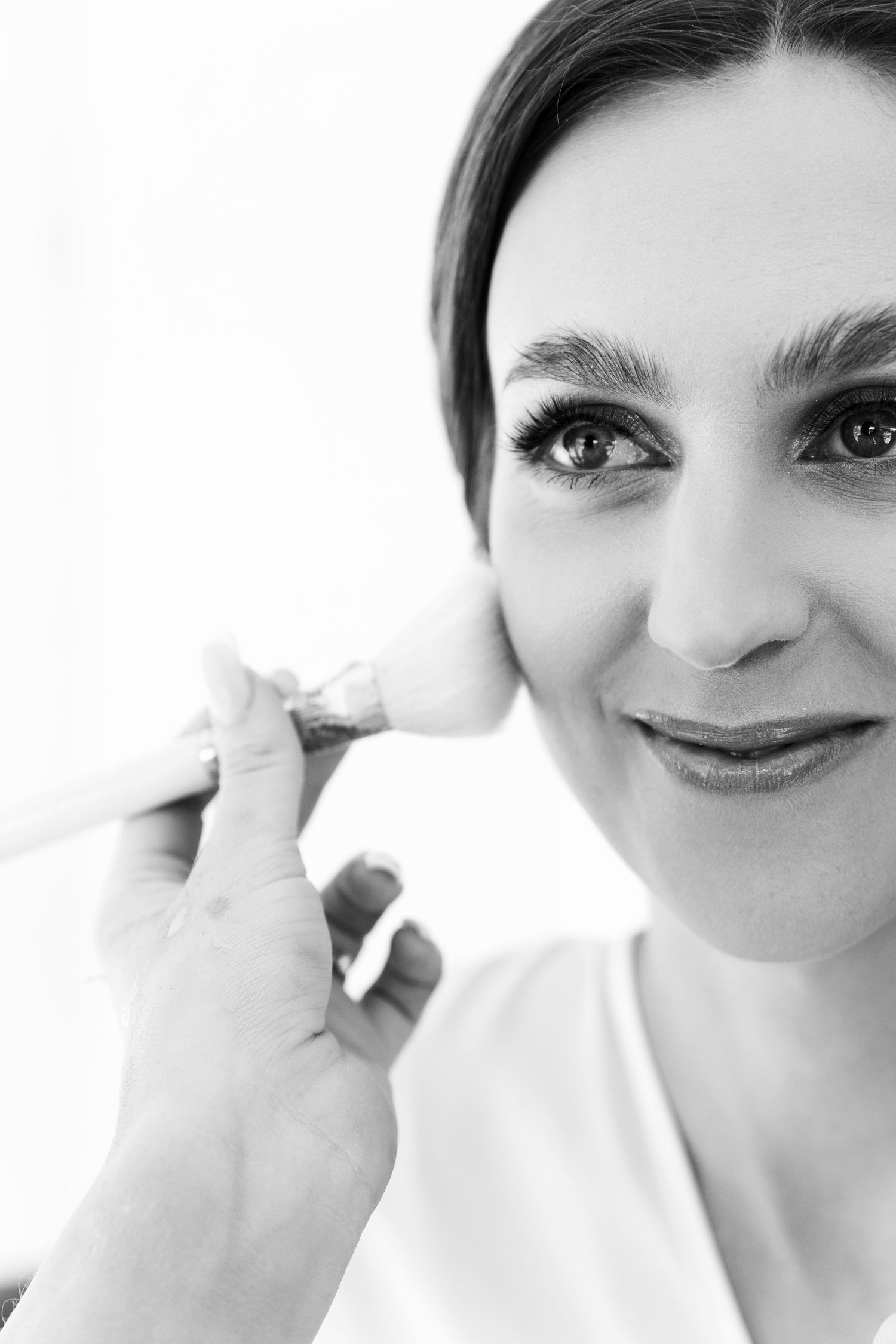 Black and white closeup portrait of someone smiling while having makeup applied with a white background.