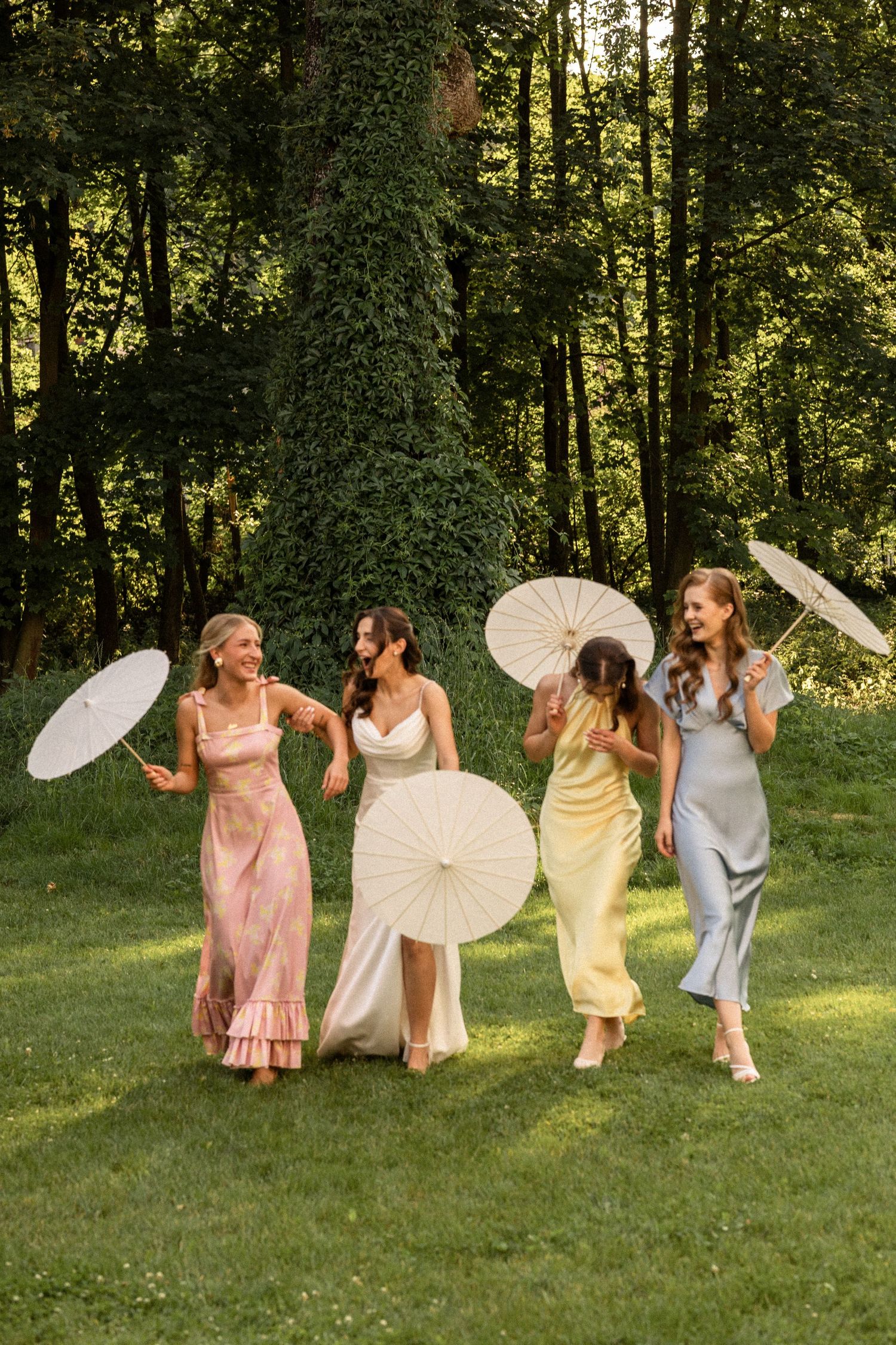 Group of friends in pastel dresses walking on grass while holding white paper parasols during a summer celebration.