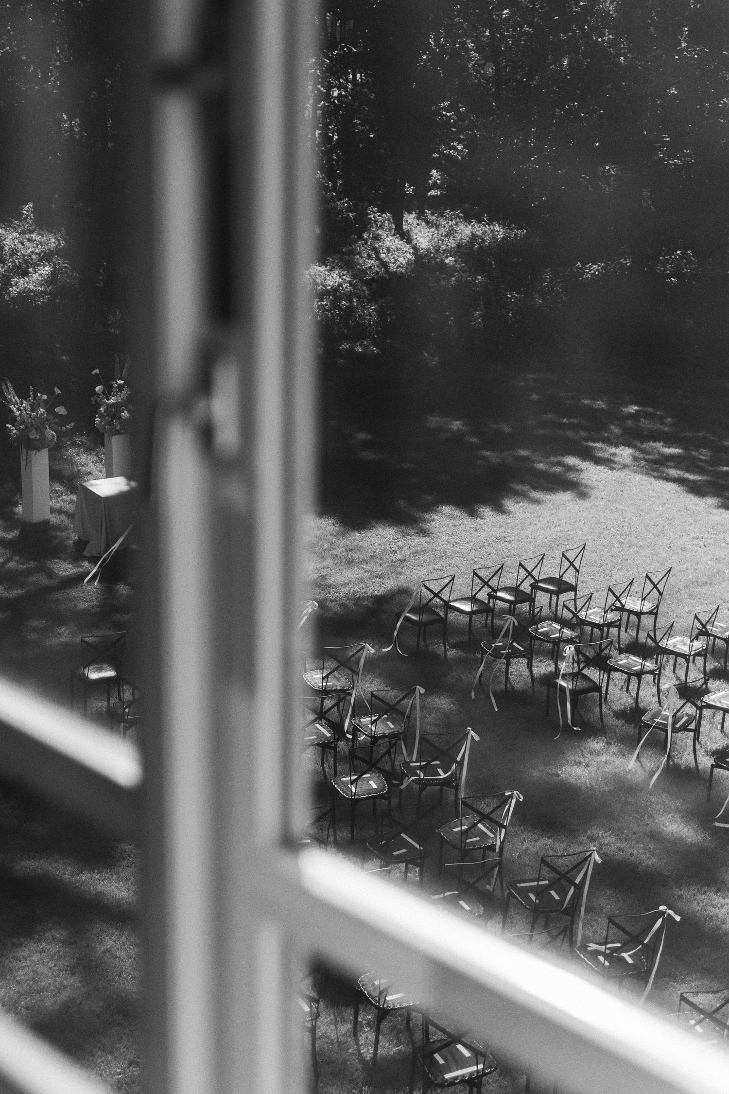 A grainy black and white image shows stacked chairs in an outdoor courtyard or patio area viewed through a window frame.
