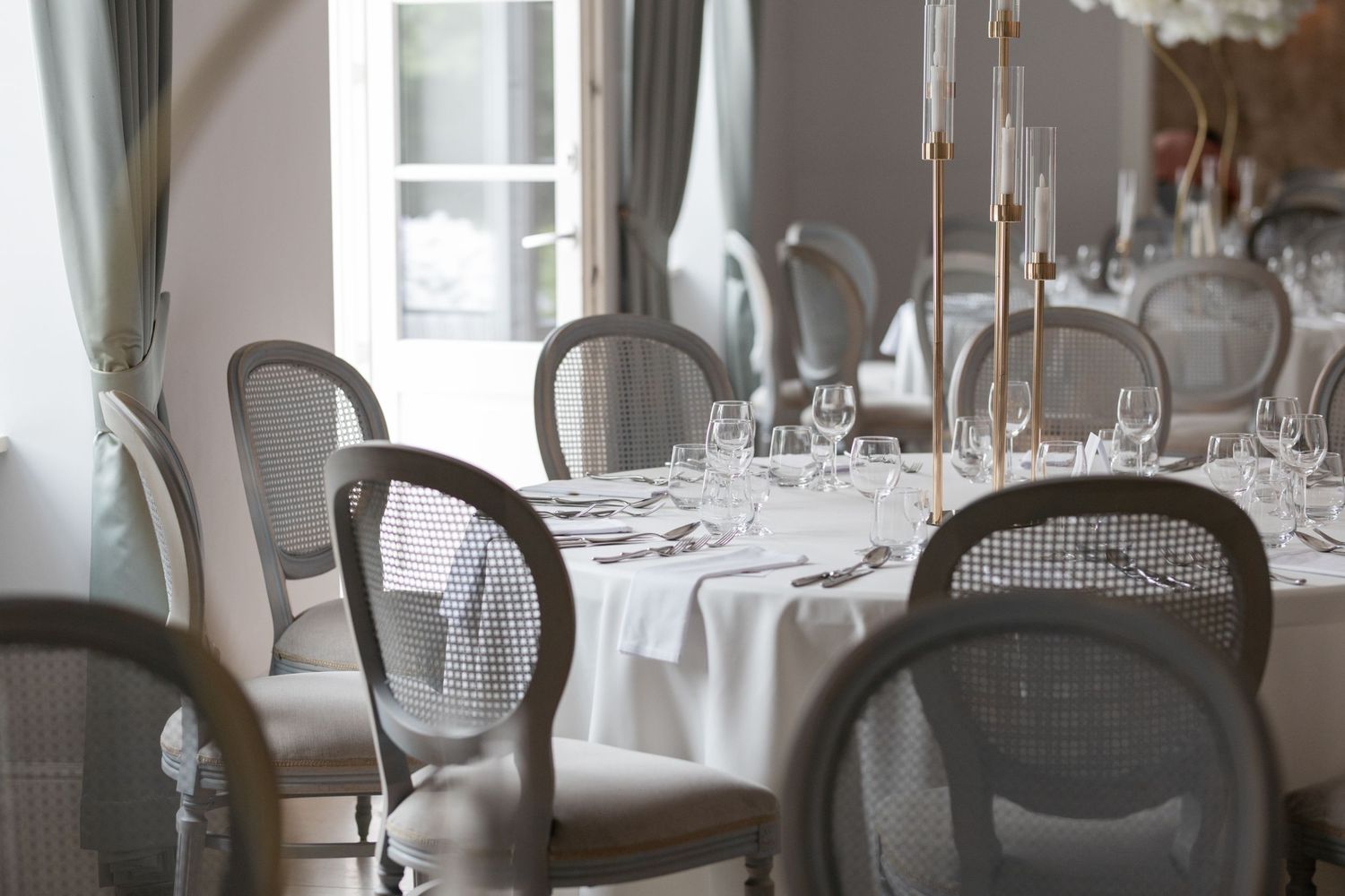 Elegant dining room with white tablecloth, crystal glasses, candelabras, and French-style cane back chairs in a bright setting.